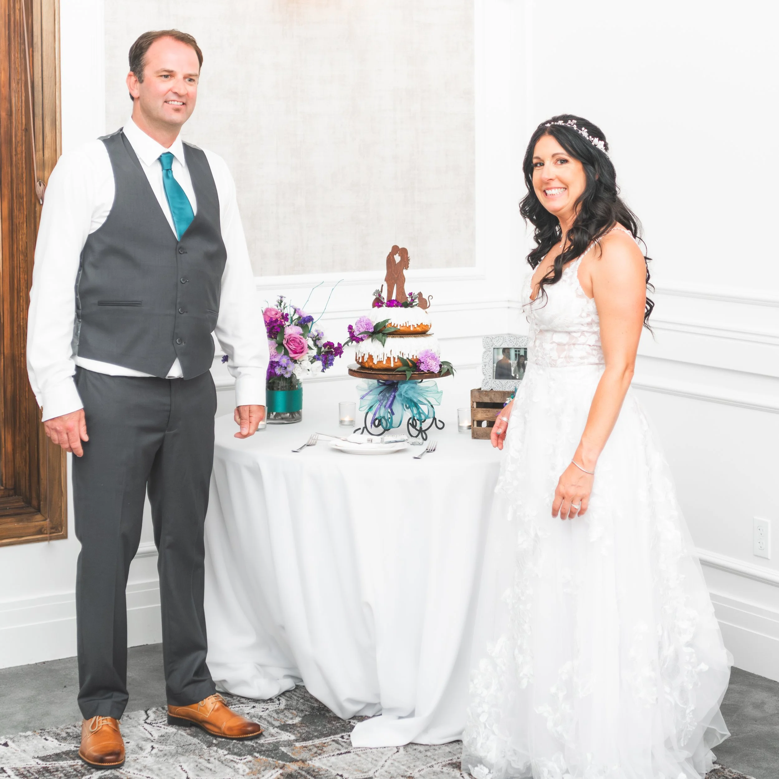 Bride with long dark hair in a white wedding dress smiling beside groom in a grey vest, white shirt, and blue tie, standing next to wedding cake decorated with purple and pink flowers.