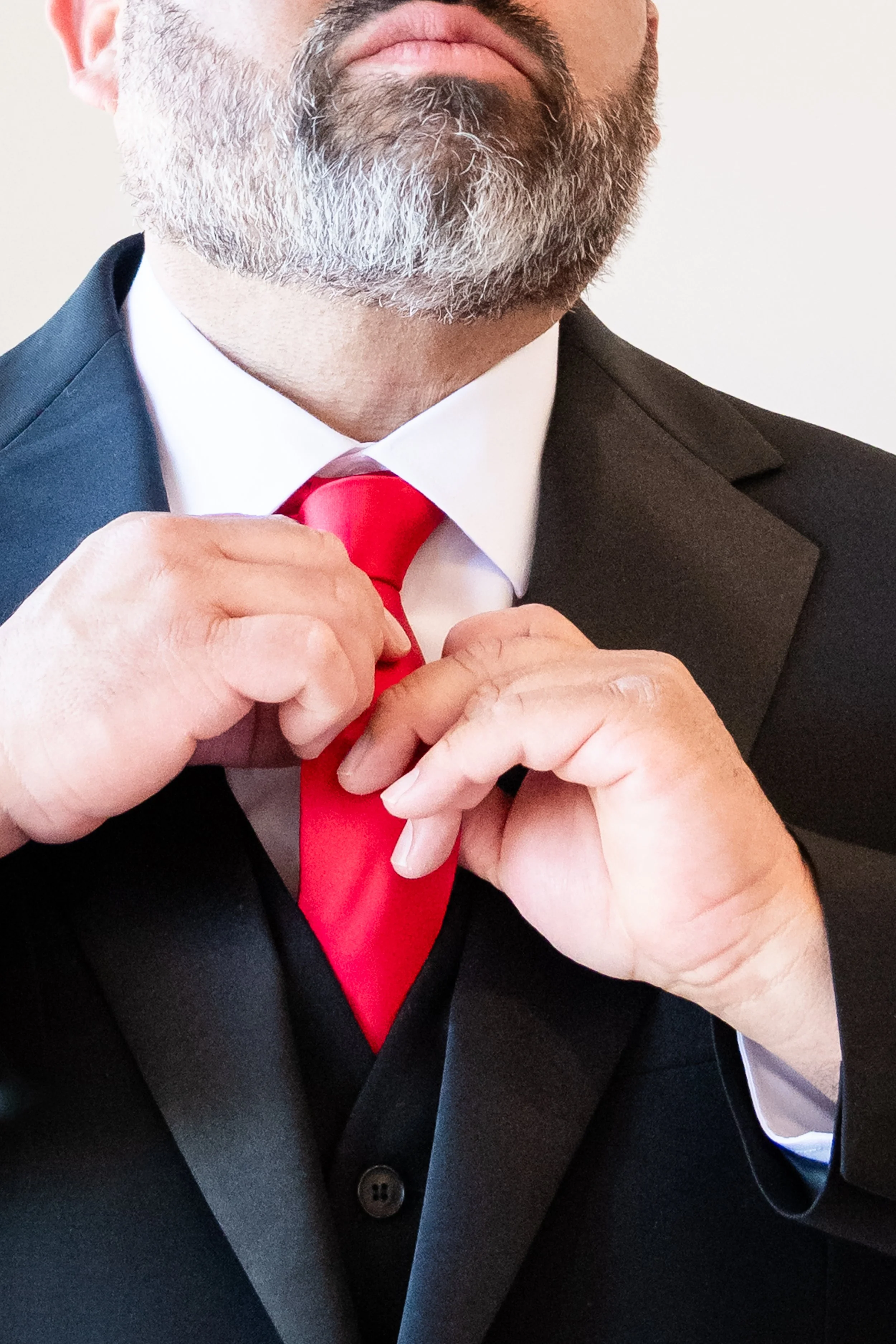 Close-up of a man adjusting a red tie, wearing a dark suit, white shirt, and fringe beard and mustache.