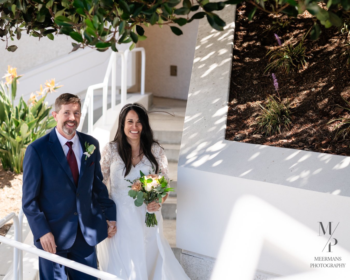 A bride and groom standing outside, smiling. The groom is wearing a blue suit with a red tie, and the bride is wearing a white wedding dress holding a bouquet of flowers. There are plants and stairs in the background.