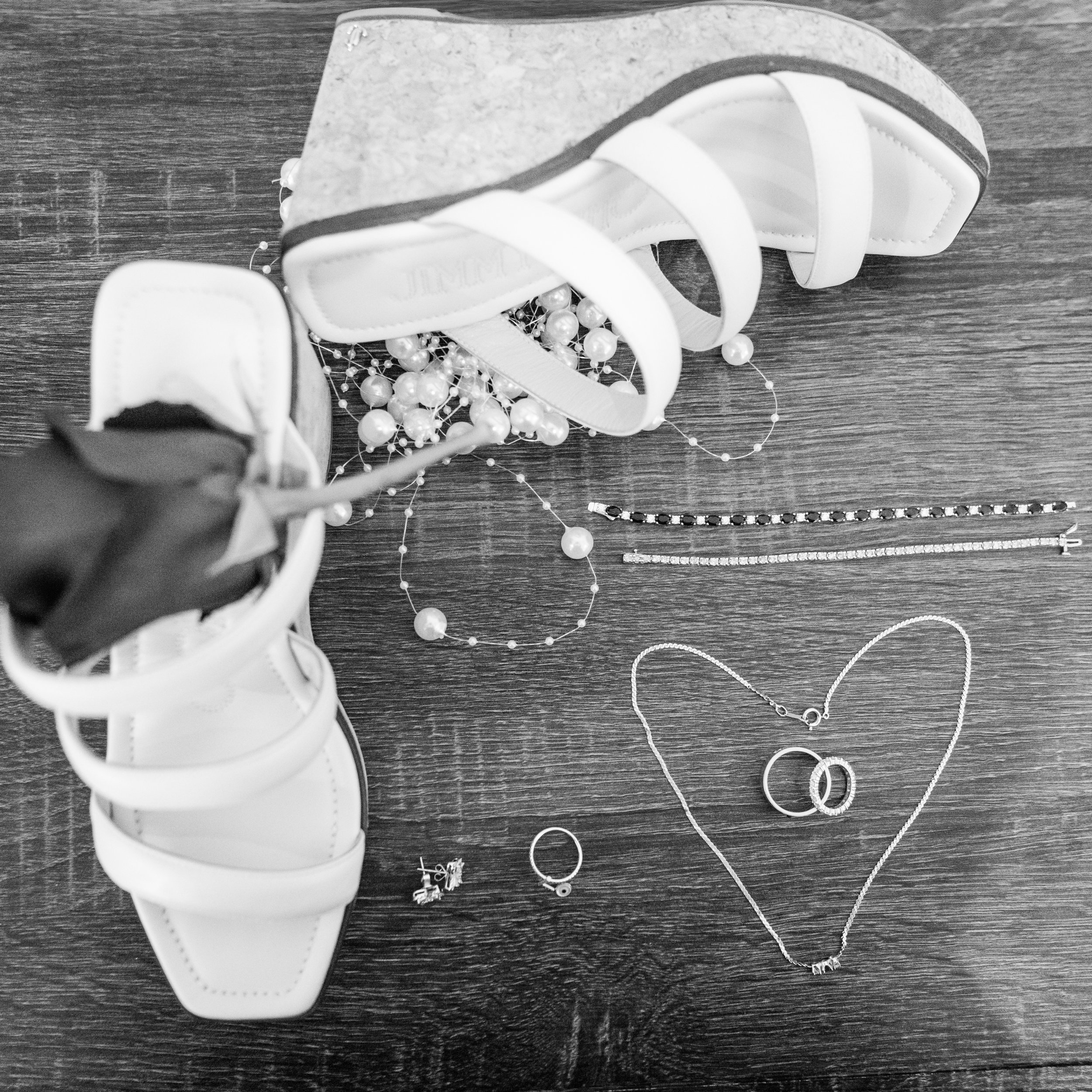 Black and white photo of a white high-heeled sandal shoe, a pearl necklace, and other jewelry pieces on a dark textured surface.