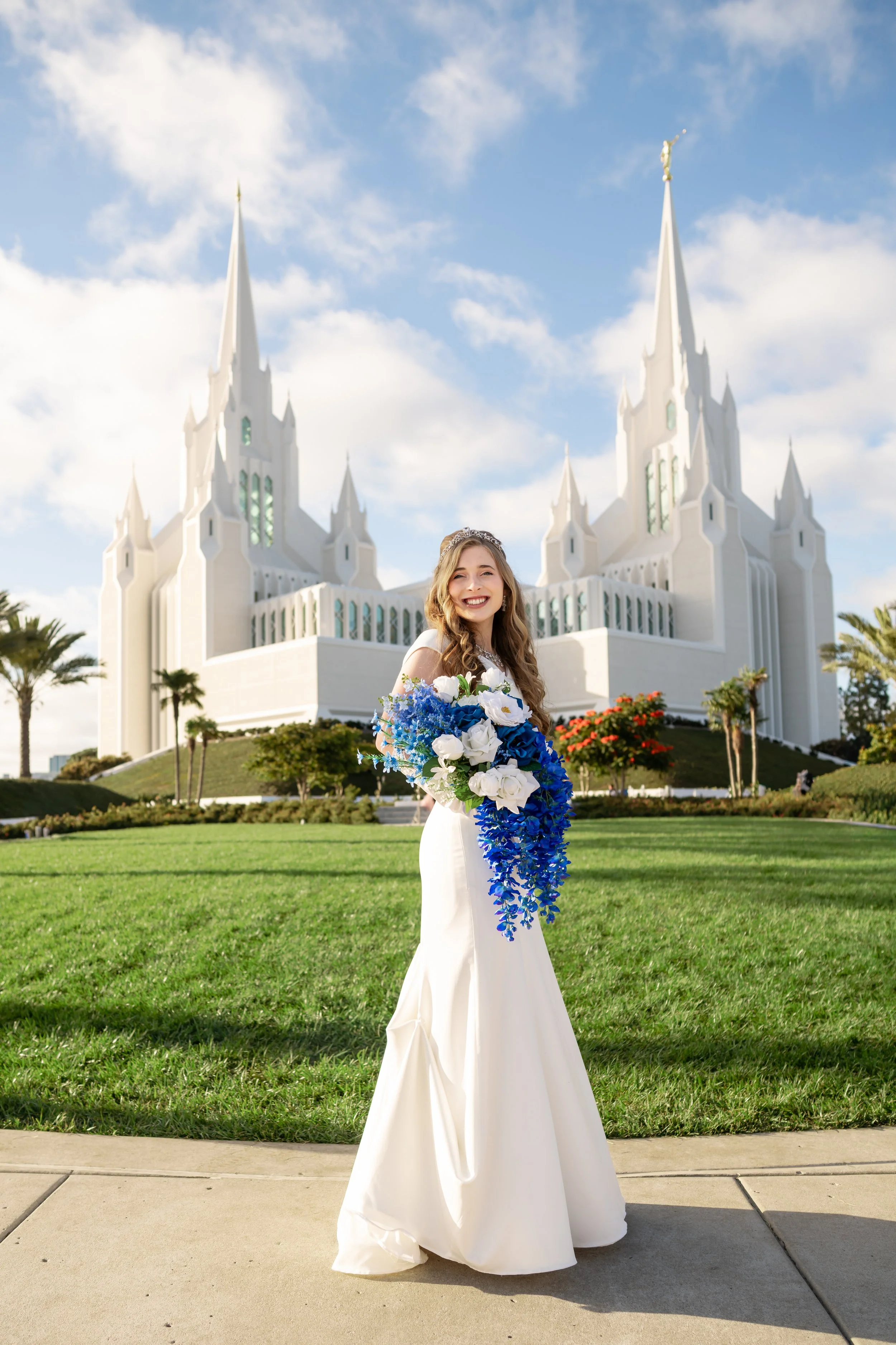 A woman in a white wedding gown holding a bouquet of blue and white flowers, standing on a sidewalk in front of a grassy landscaped area with trees and a large white church featuring multiple spires in the background, under a partly cloudy sky.