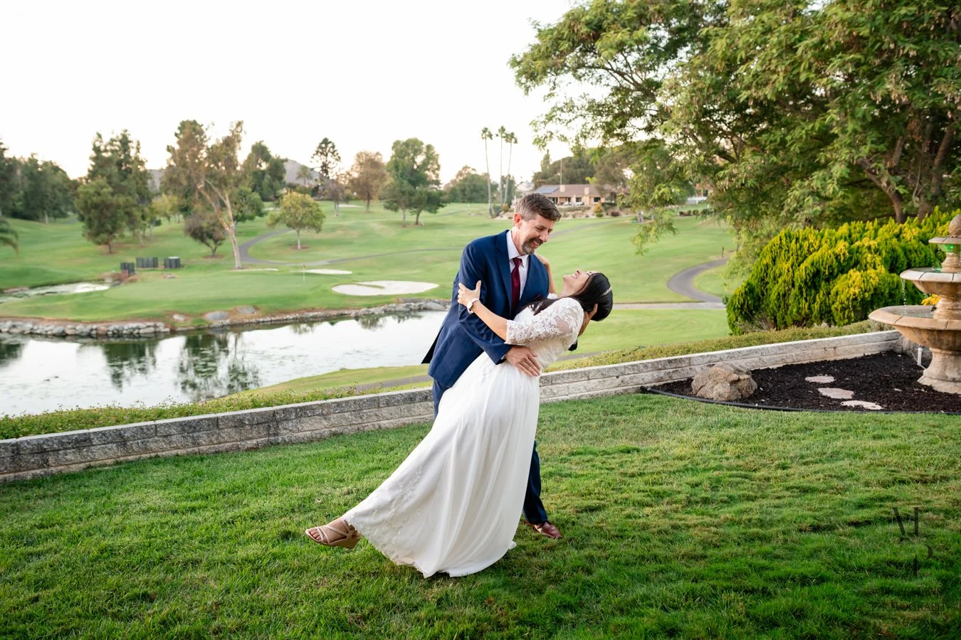 A happy couple, dressed in wedding attire, dancing outdoors on a grassy area near a pond with trees and a golf course in the background.