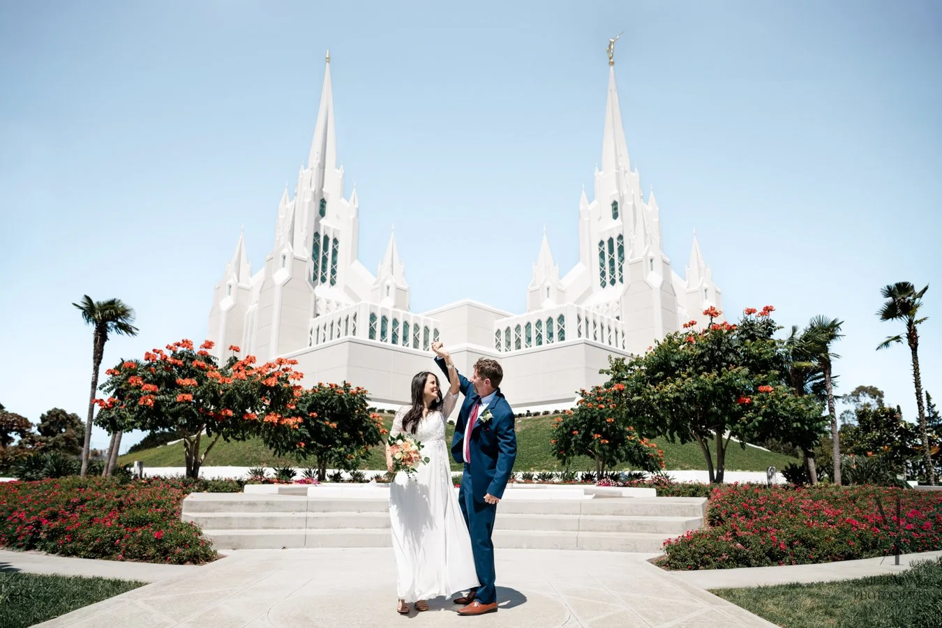 A newlywed couple in wedding attire in front of a large white church with tall spires, surrounded by palm trees and flowering bushes.