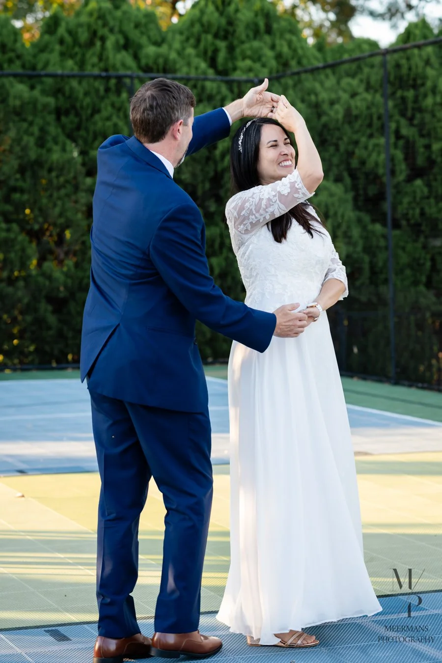 A man in a blue suit twirling a woman in a white dress on an outdoor tennis court with green trees in the background.