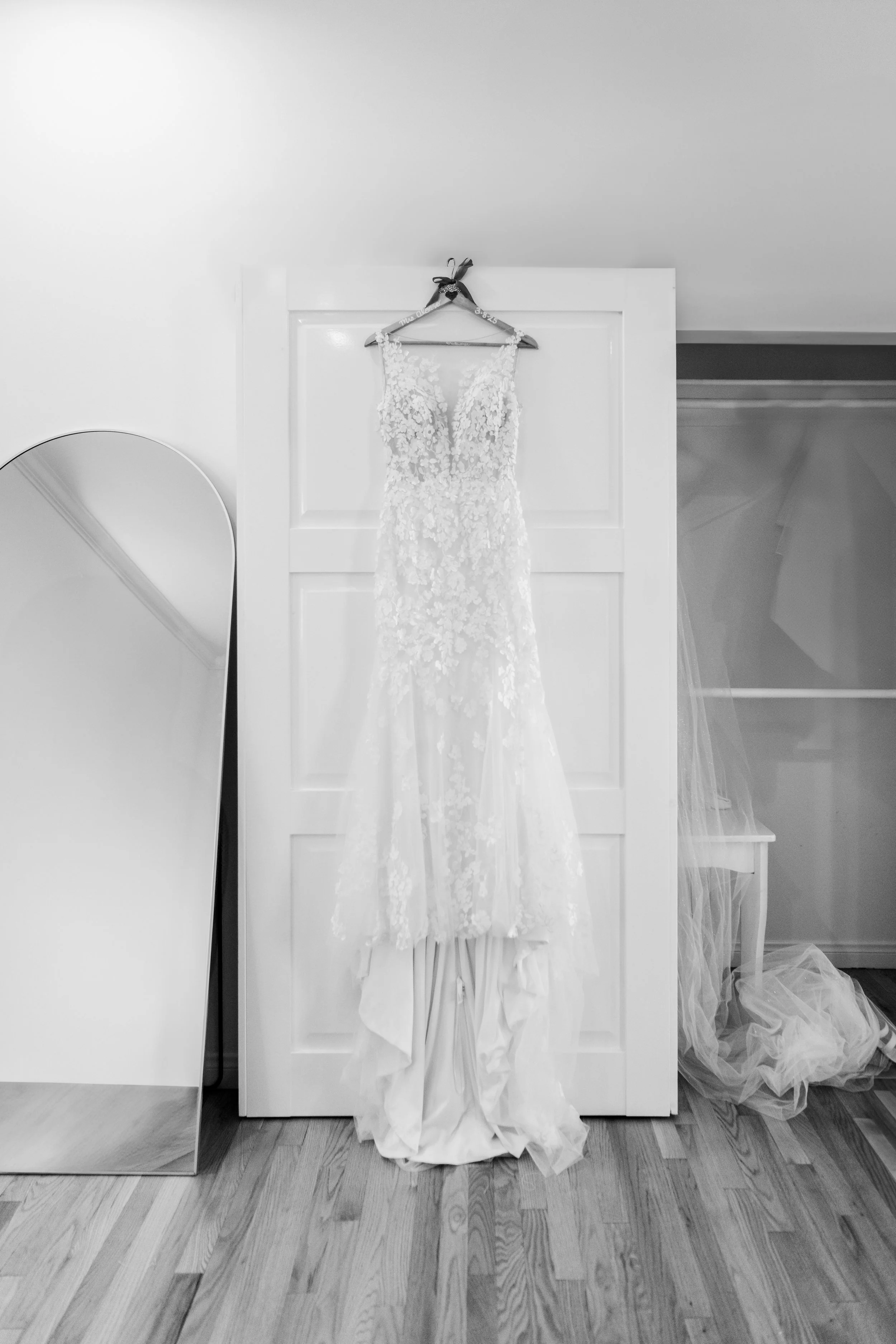 A wedding dress hangs on a hanger on top of a white door. The dress has lace and floral embroidery details and a long train hanging down. There is a full-length mirror to the left and some tulle fabric on the floor.