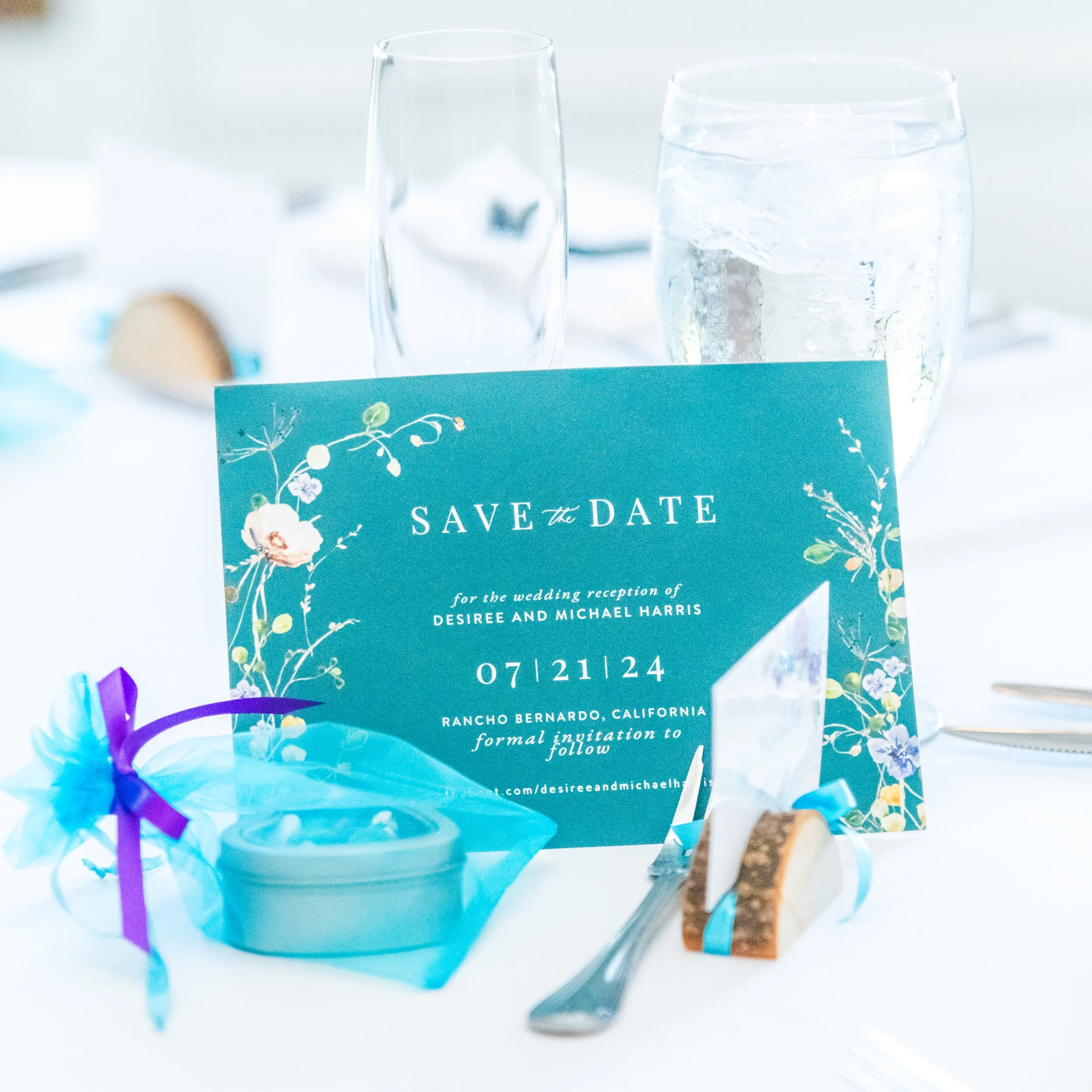 Close-up of a wedding reception table with a blue 'Save the Date' card, a glass of water with ice, and colorful wrapped candies.