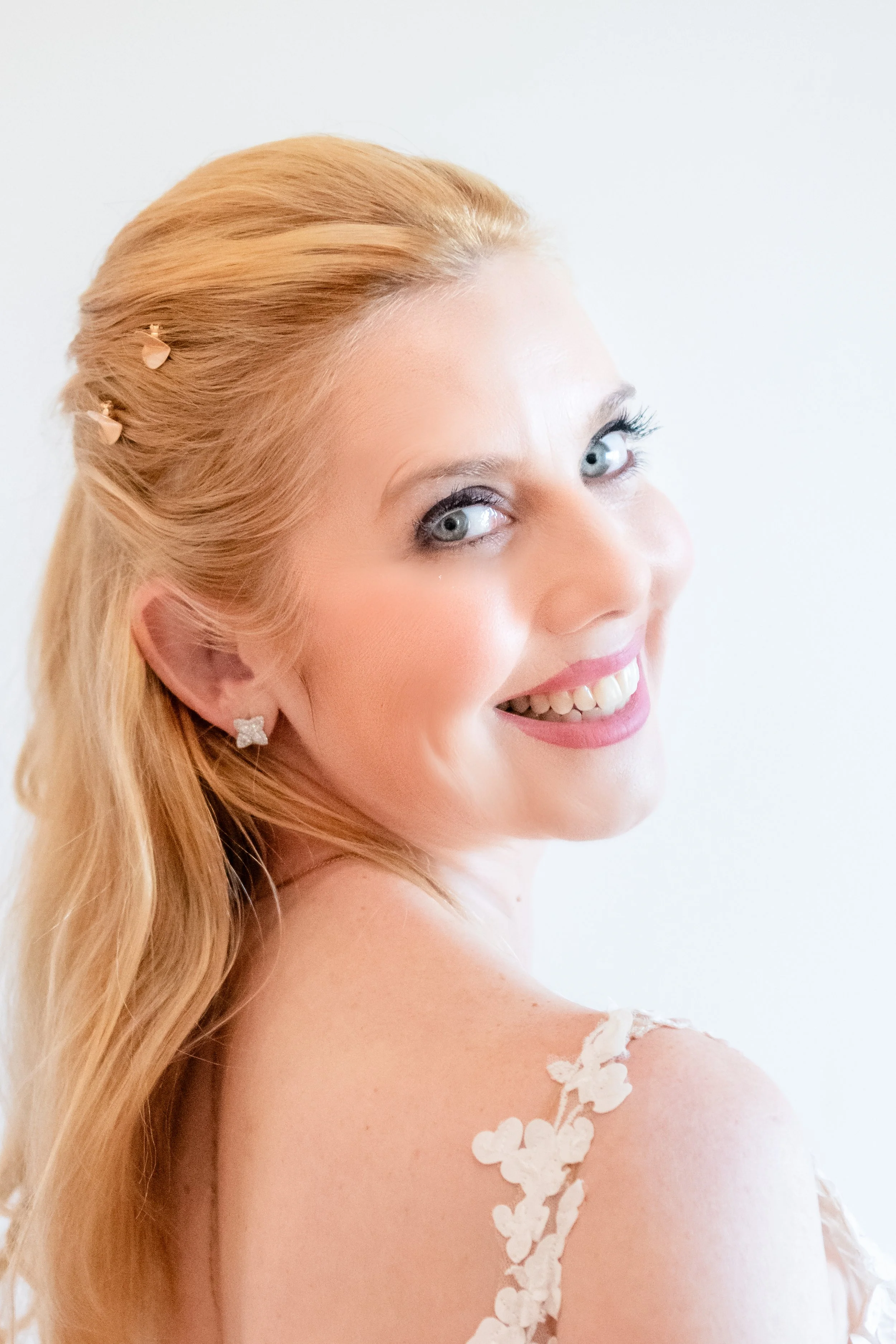 Close-up of a smiling woman with long blonde hair, blue eyes, and butterfly-shaped earrings, wearing a white floral dress, posing against a plain white background.