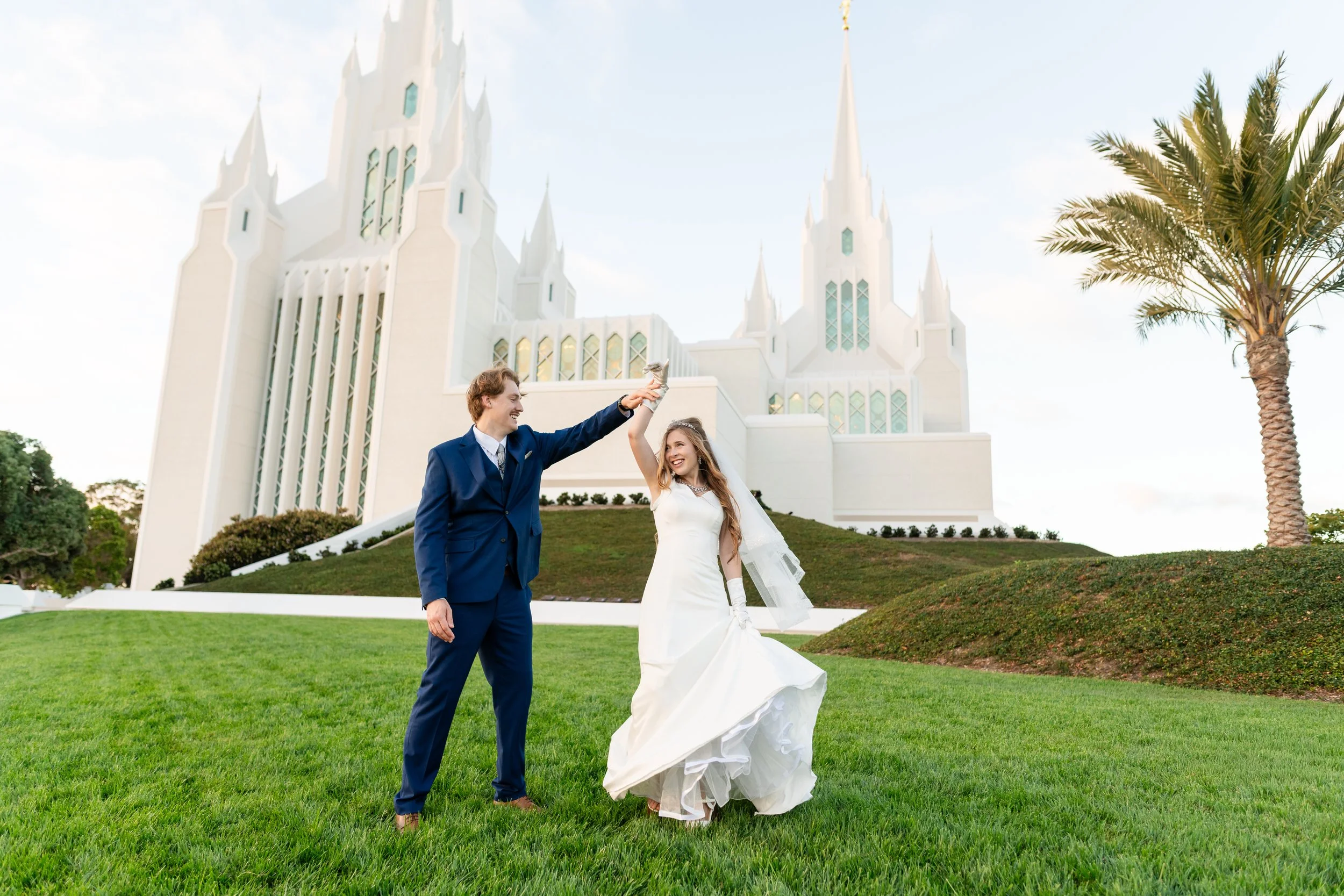 A newlywed couple dancing on a grassy lawn in front of a white church with tall spires, a palm tree on the right, and clear blue sky.