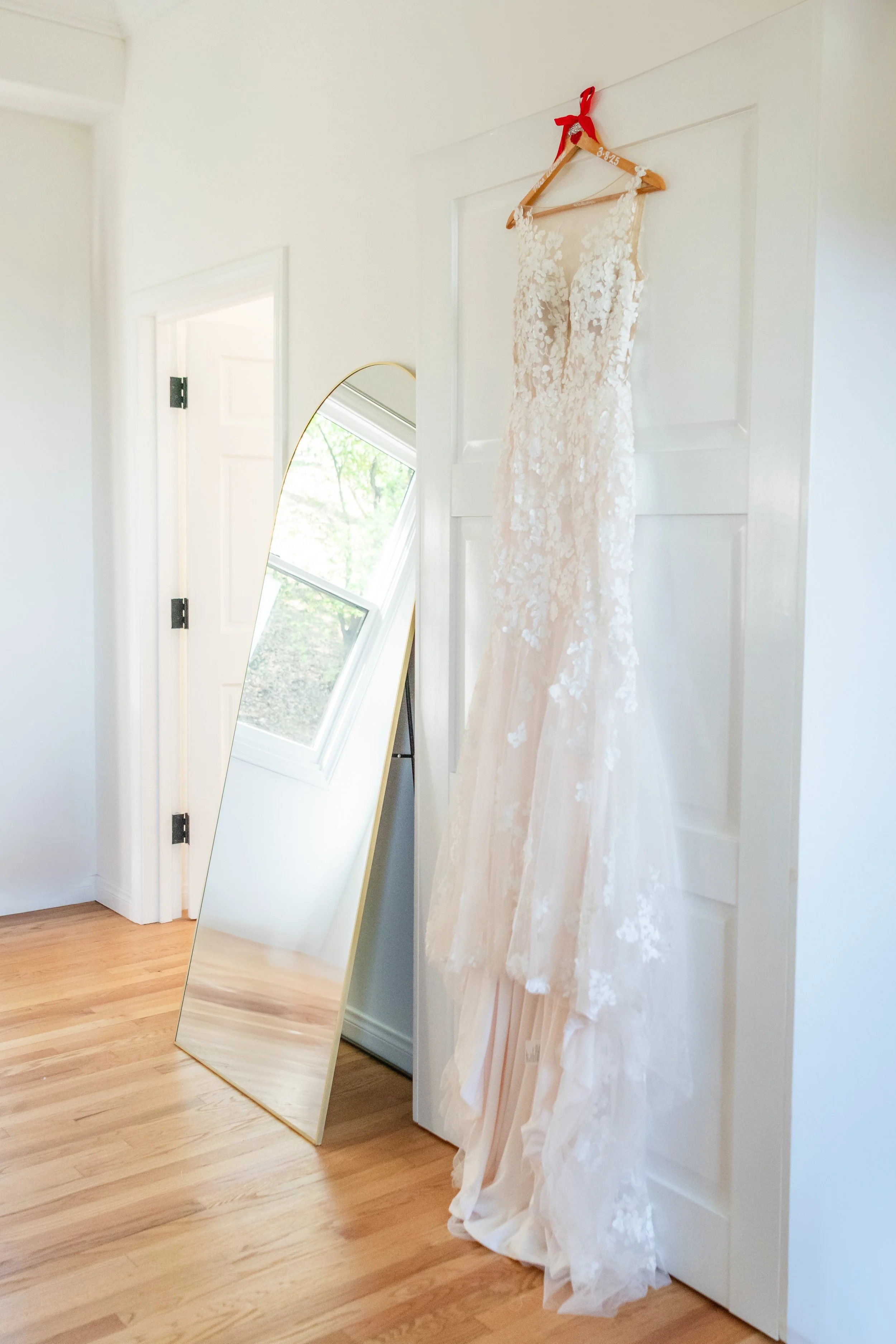 A wedding dress hanging on a wooden hanger with a red ribbon, next to a full-length mirror leaning against a white door inside a room with hardwood floors.