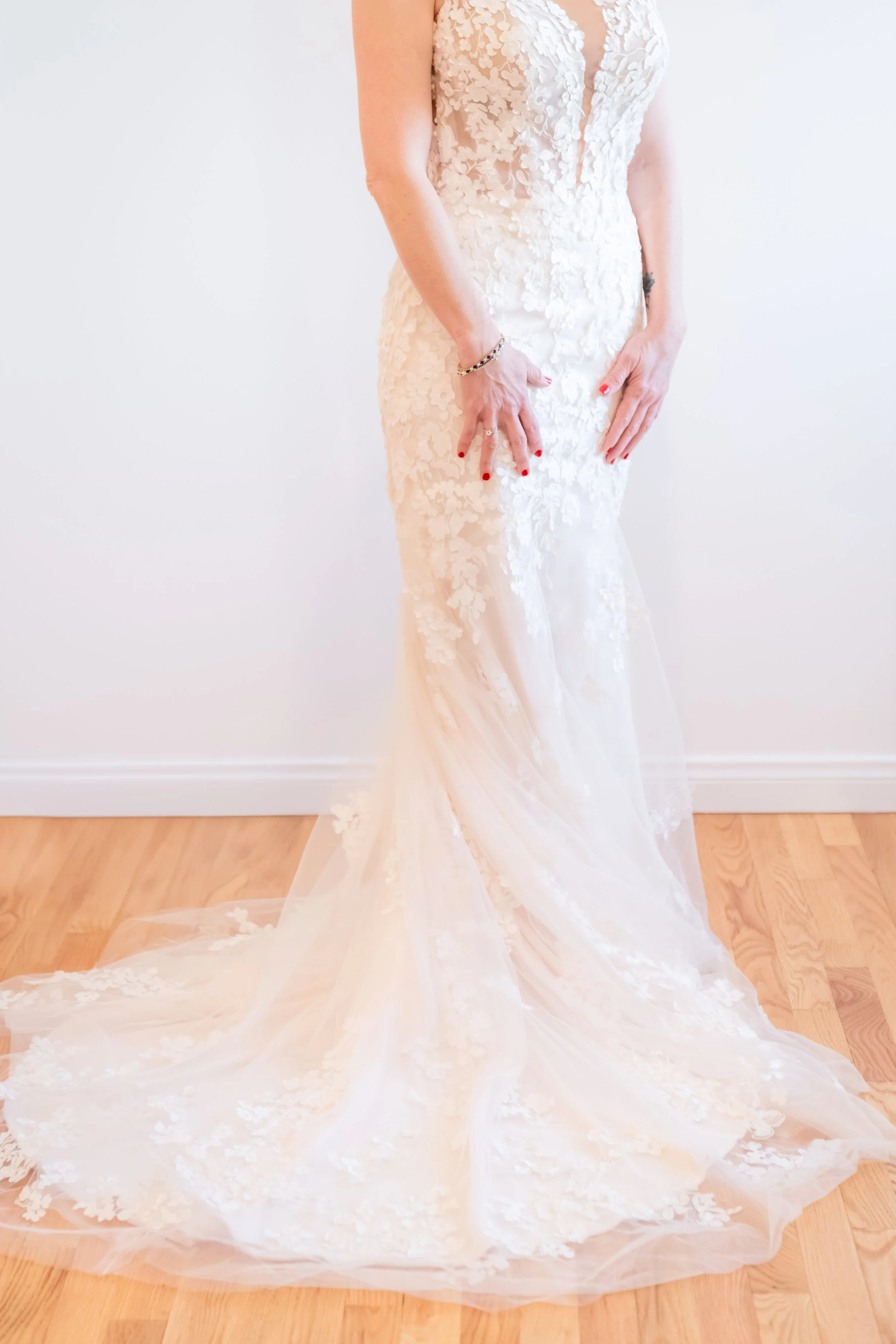 A bride is wearing a white wedding dress with floral lace details, a deep V neckline, and a long flowing train. She is standing on a wooden floor with a plain white wall background, showcasing her dress and jewelry.