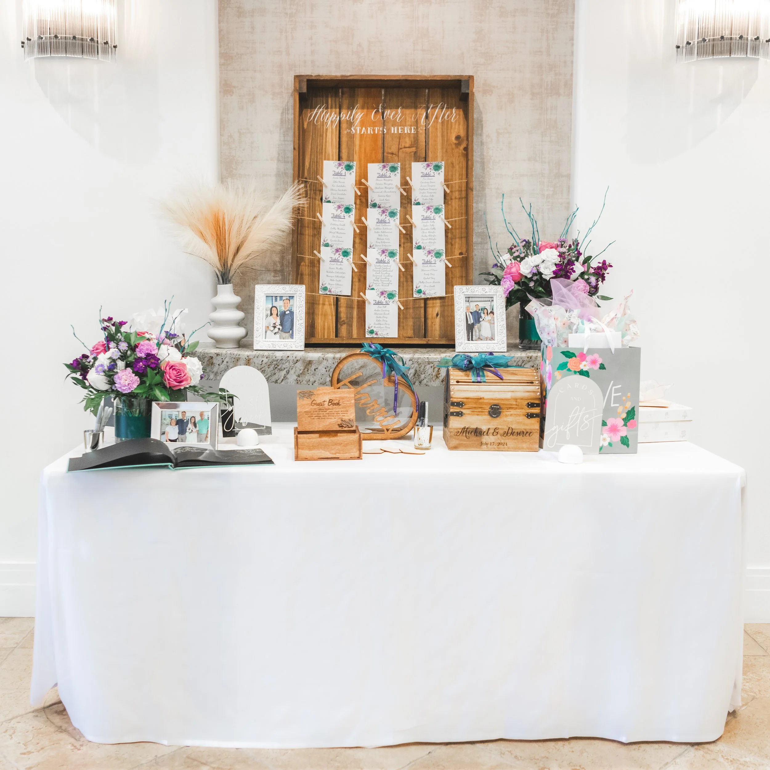 Wedding reception table with floral arrangements, framed photos of the couple, guest book, and decorative gift boxes, set against a backdrop with wooden sign inscription 'Happily Ever After Starts Here'.