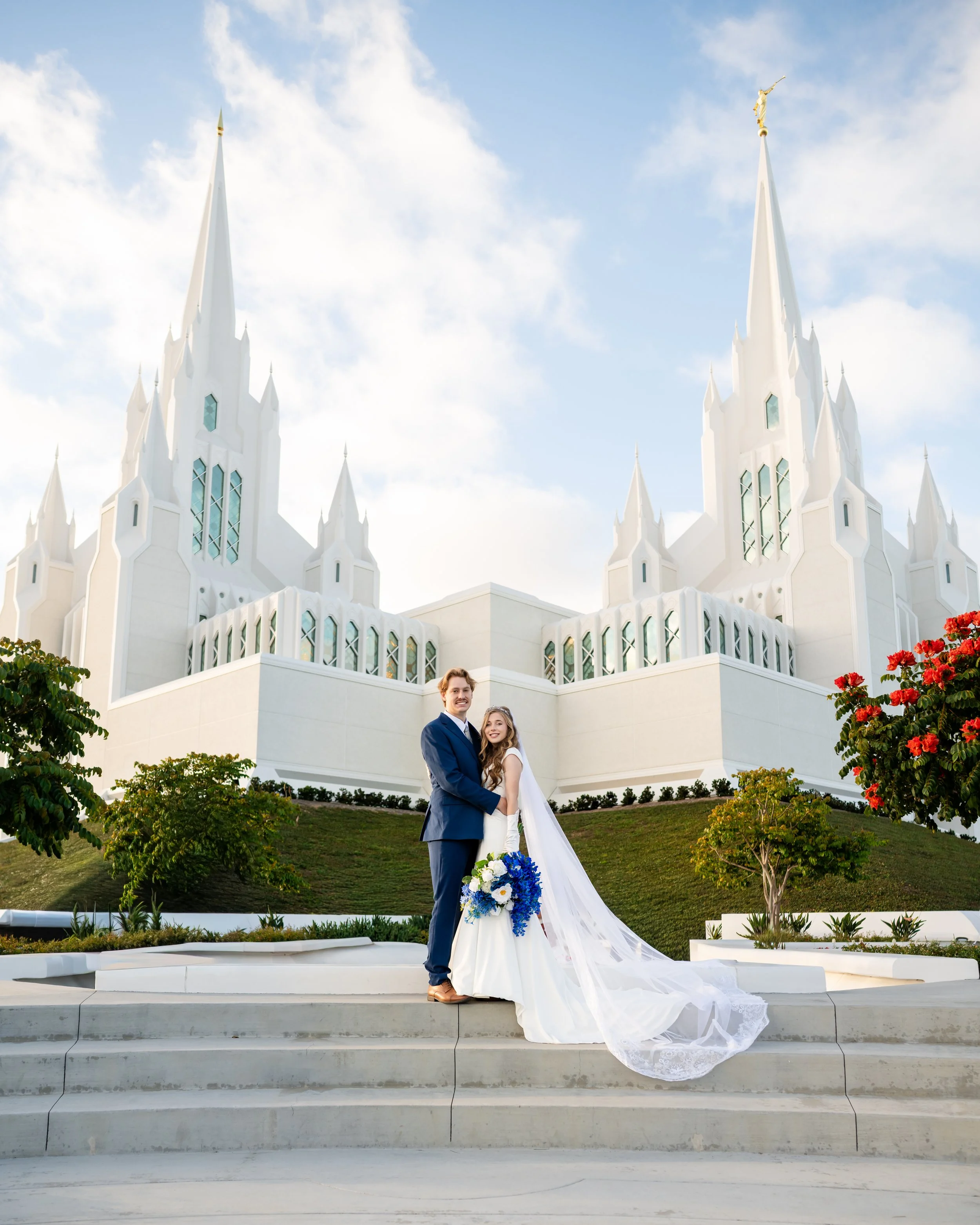 A bride and groom standing on the steps of a white church or chapel, smiling and holding each other, with greenery and red flowers in the foreground and a partly cloudy sky above.