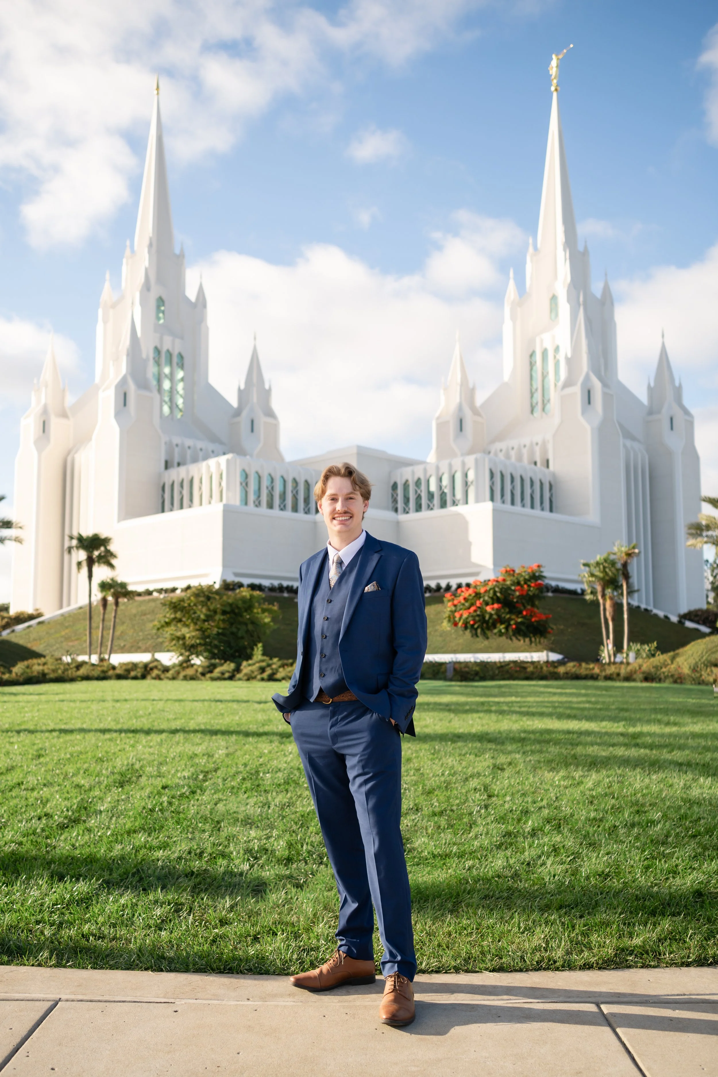 A man in a blue suit standing outdoors on a sidewalk in front of a grassy area, with the Salt Lake Temple in the background under a blue sky with scattered clouds.