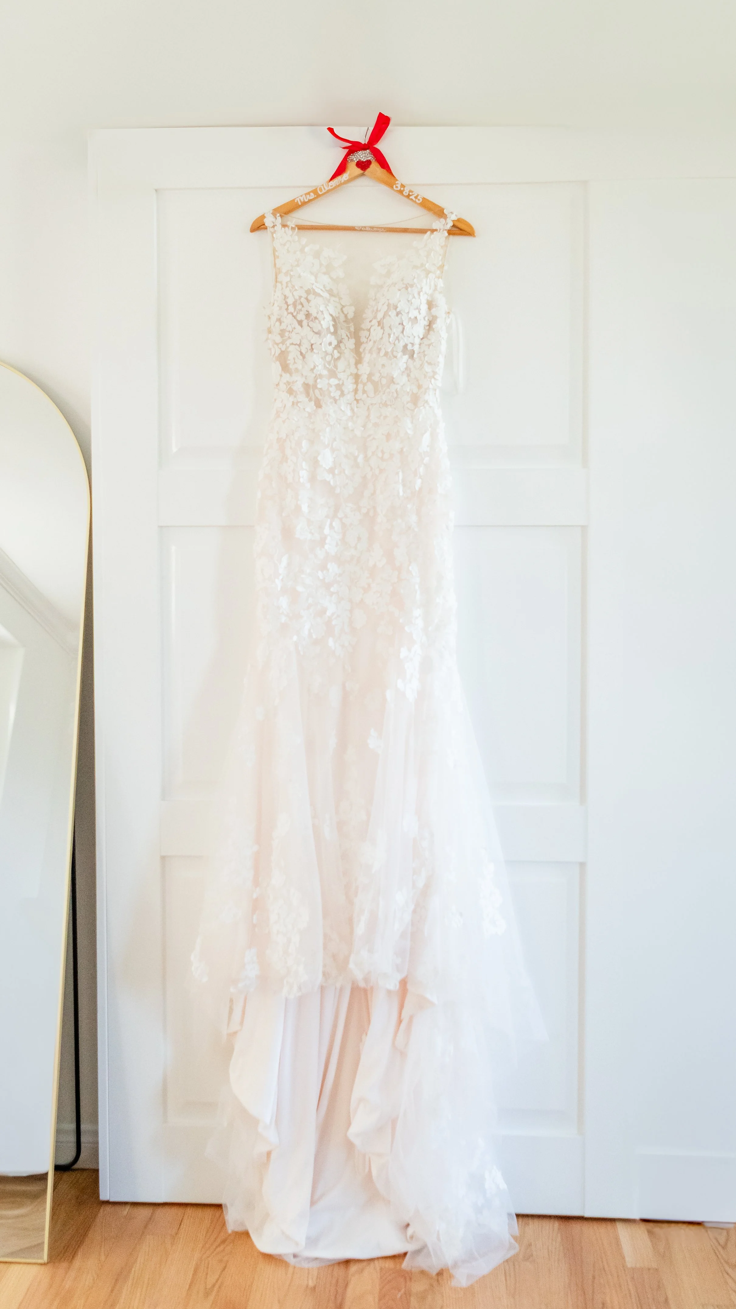 A white lace wedding dress hanging on a wooden hanger with a red ribbon on a white door.