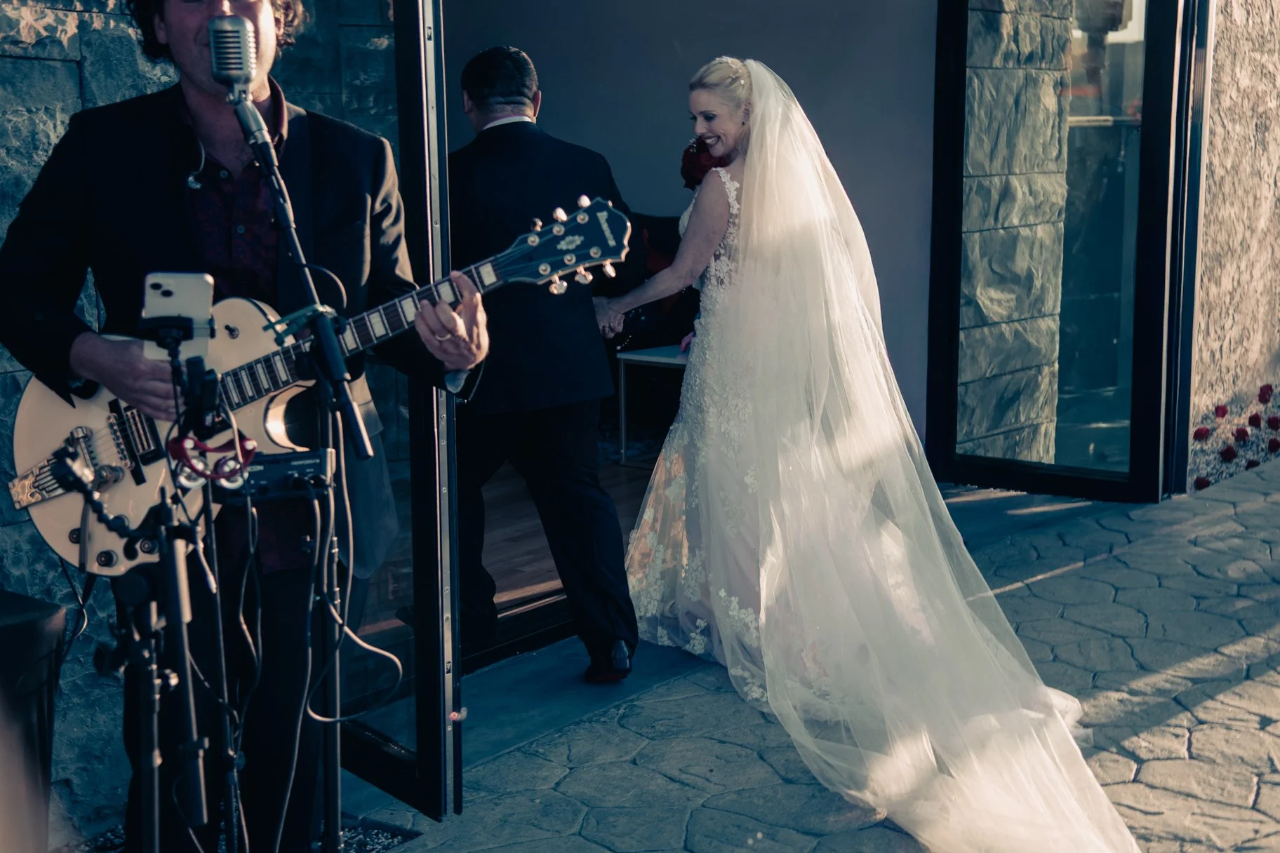 Bride in a white wedding dress holding hands with groom in a black suit, inside a building with stone walls, as a musician plays guitar nearby.