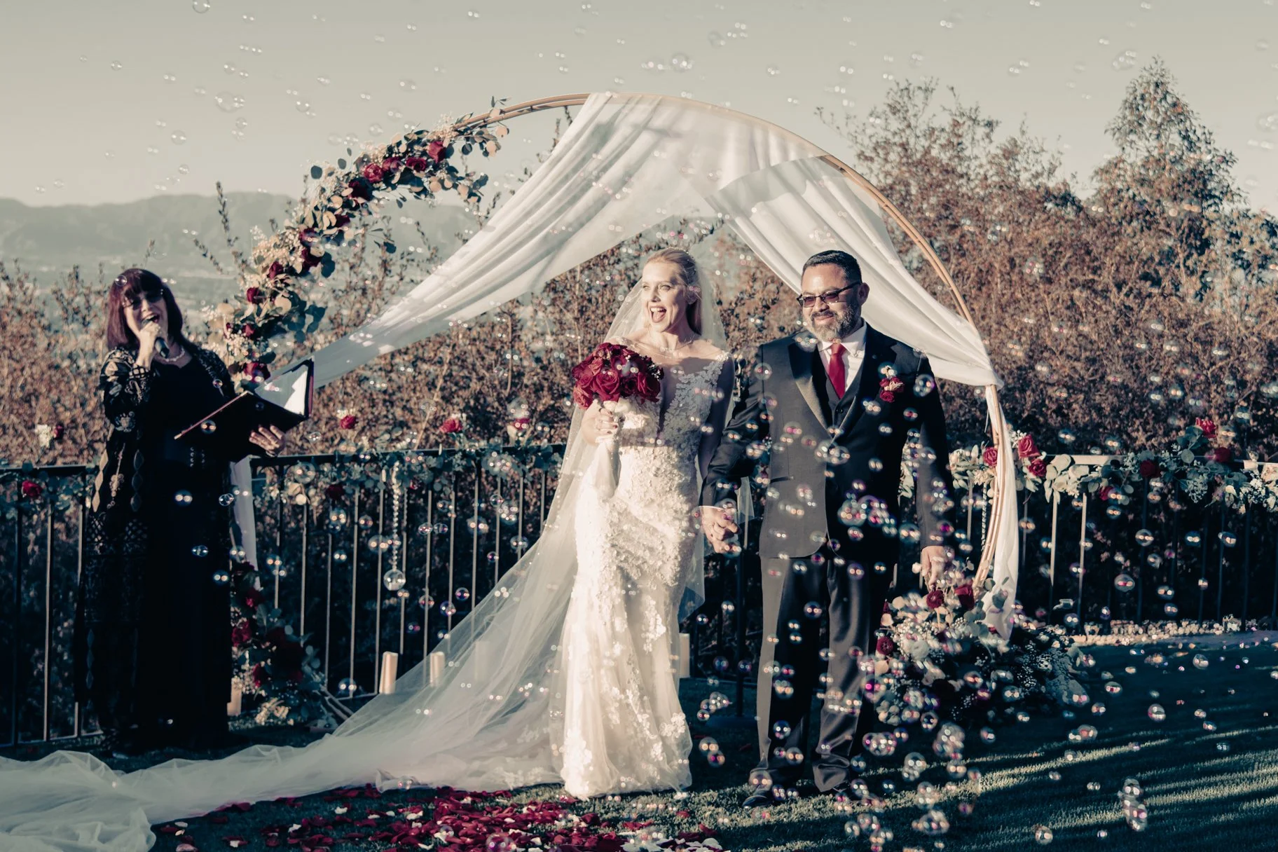 A bride and groom hold hands and smile while walking down an outdoor wedding aisle decorated with flowers and surrounded by falling flower petals or confetti, under a floral and fabric-covered arch.