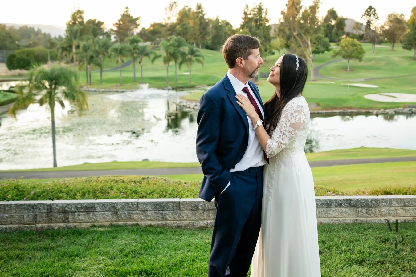 A bride and groom stand close together on a golf course, looking into each other's eyes, with water and trees in the background during sunset.