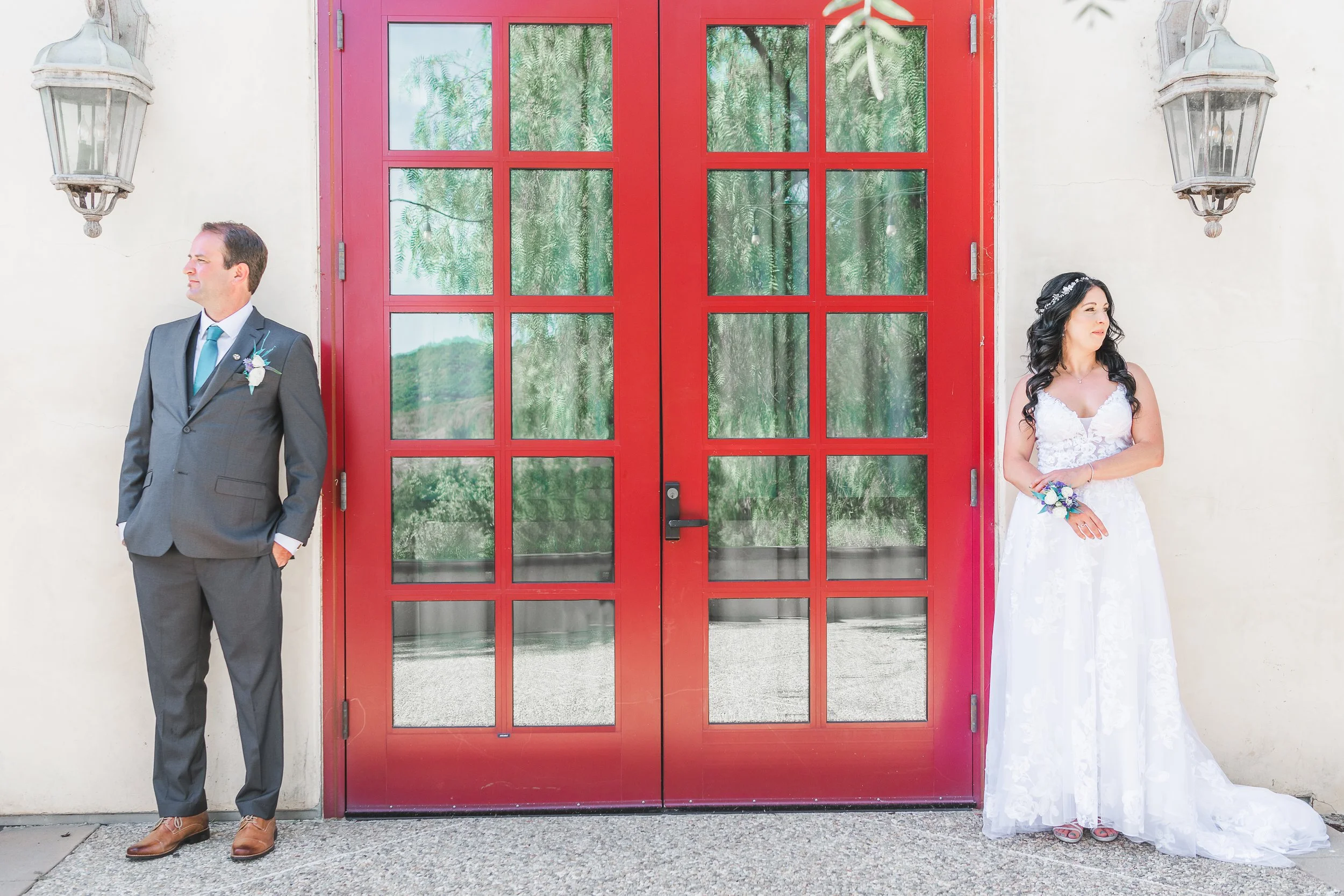 A bride and groom stand on either side of a red door with glass panels, with the groom on the left and the bride on the right, holding a bouquet, both looking away from each other.