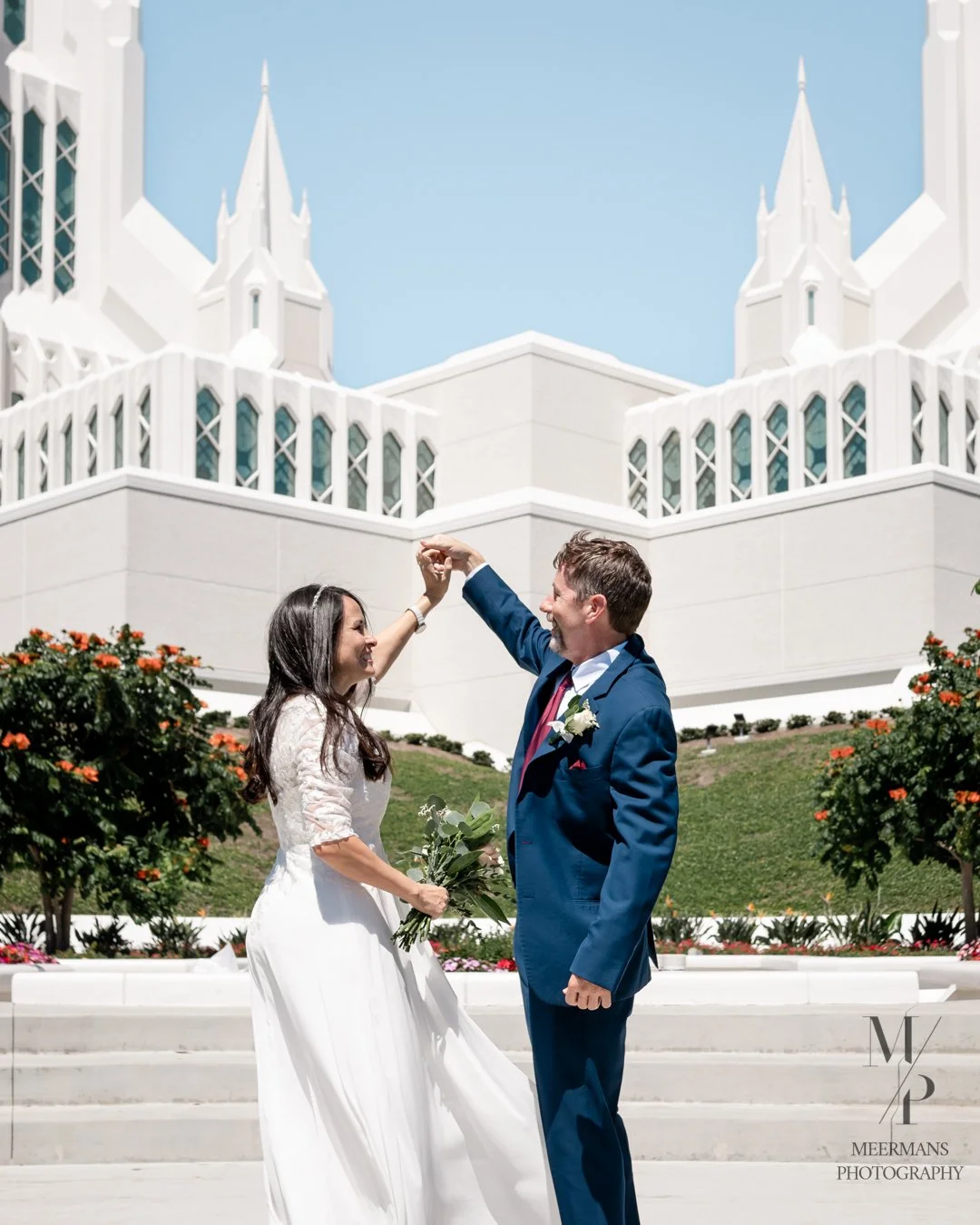 A newly married couple dancing outdoors in front of a white architectural building, with the woman holding a bouquet and the man in a blue suit, smiling at each other.