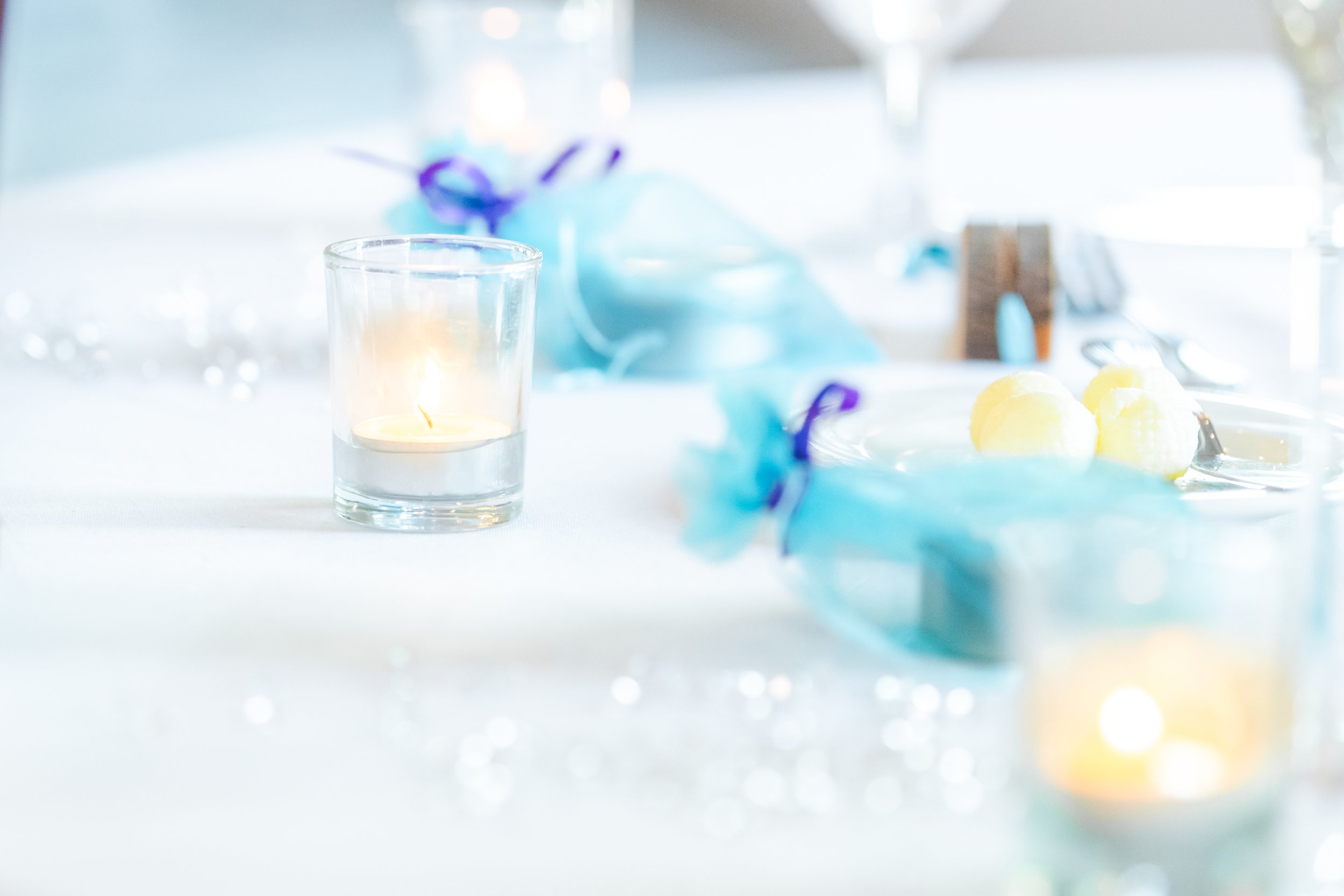 A lit candle in a transparent glass holder, blurred blue decorations, and yellow objects on a white table.
