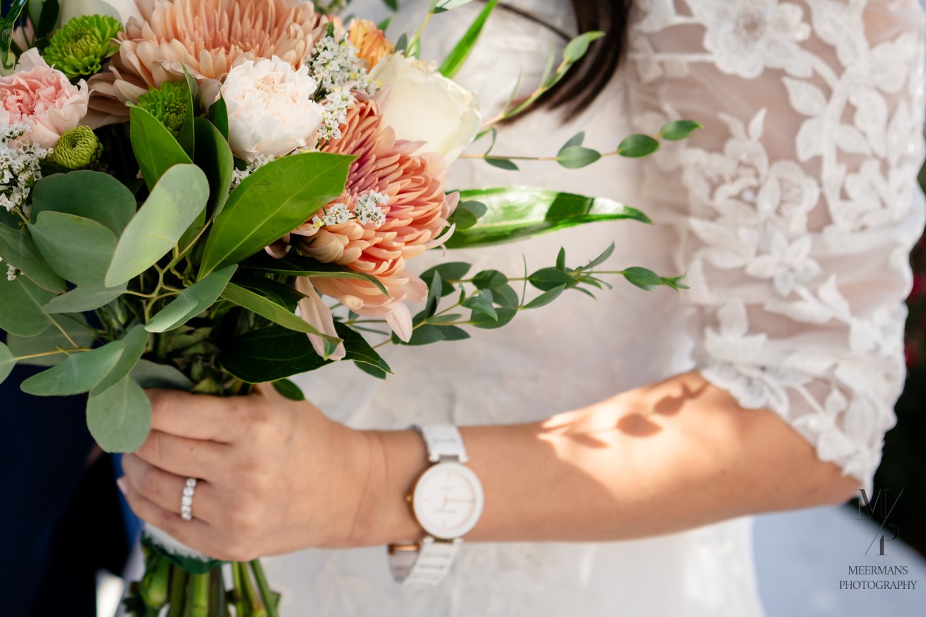 A bride holding a bouquet of pink, peach, white flowers and greenery, wearing a white lace wedding dress, with a wristwatch and wedding ring visible.