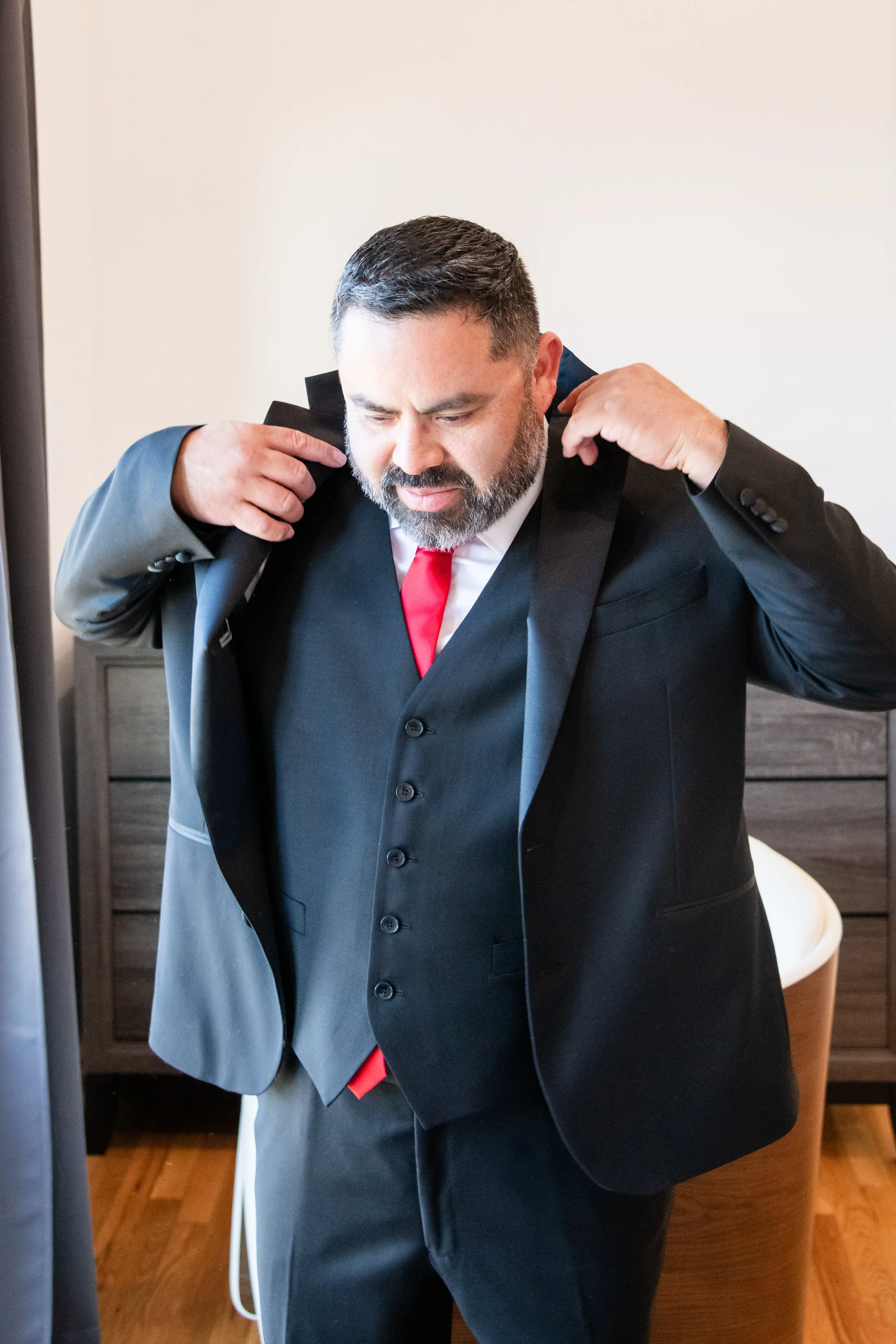 A man with dark hair and a beard is adjusting his black suit jacket with a white shirt and red tie. He is standing indoors near a wooden dresser, with a white chair and a beige wall in the background.