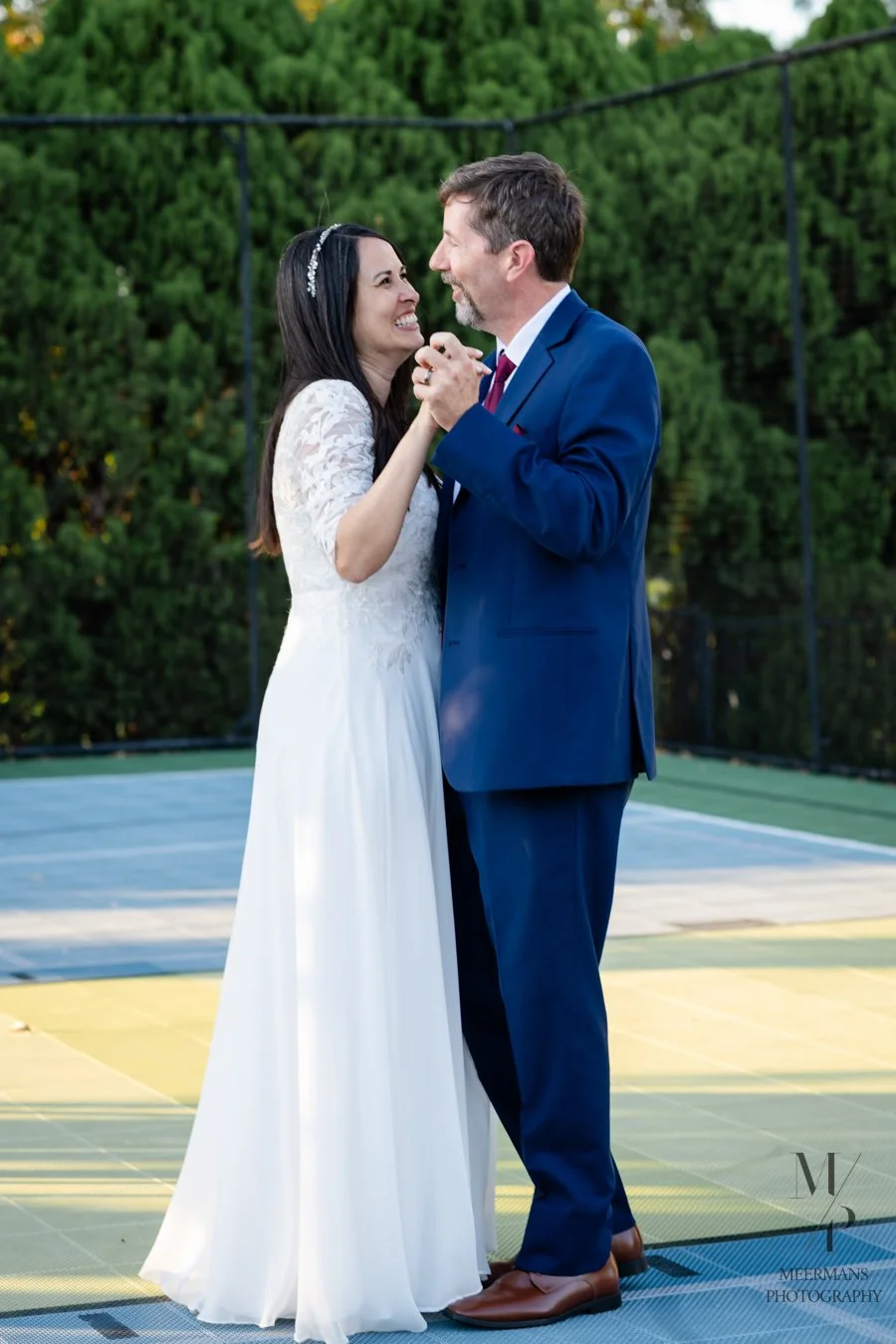 A bride and groom dancing on a court with green trees in the background, smiling and looking at each other happily.