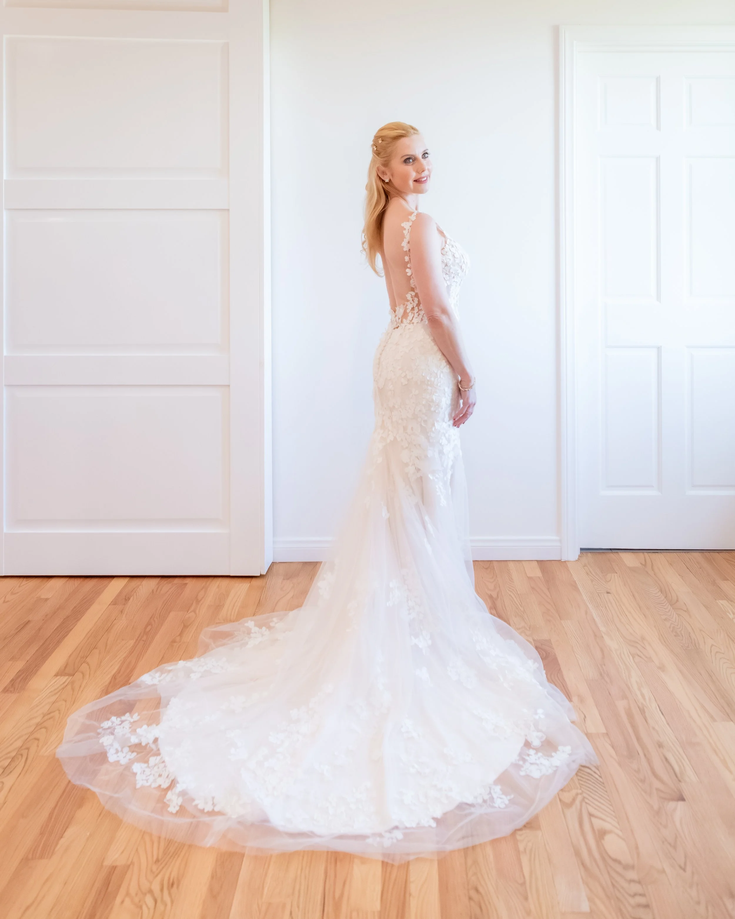 A woman in a white wedding dress standing indoors on a hardwood floor, smiling and looking over her shoulder.