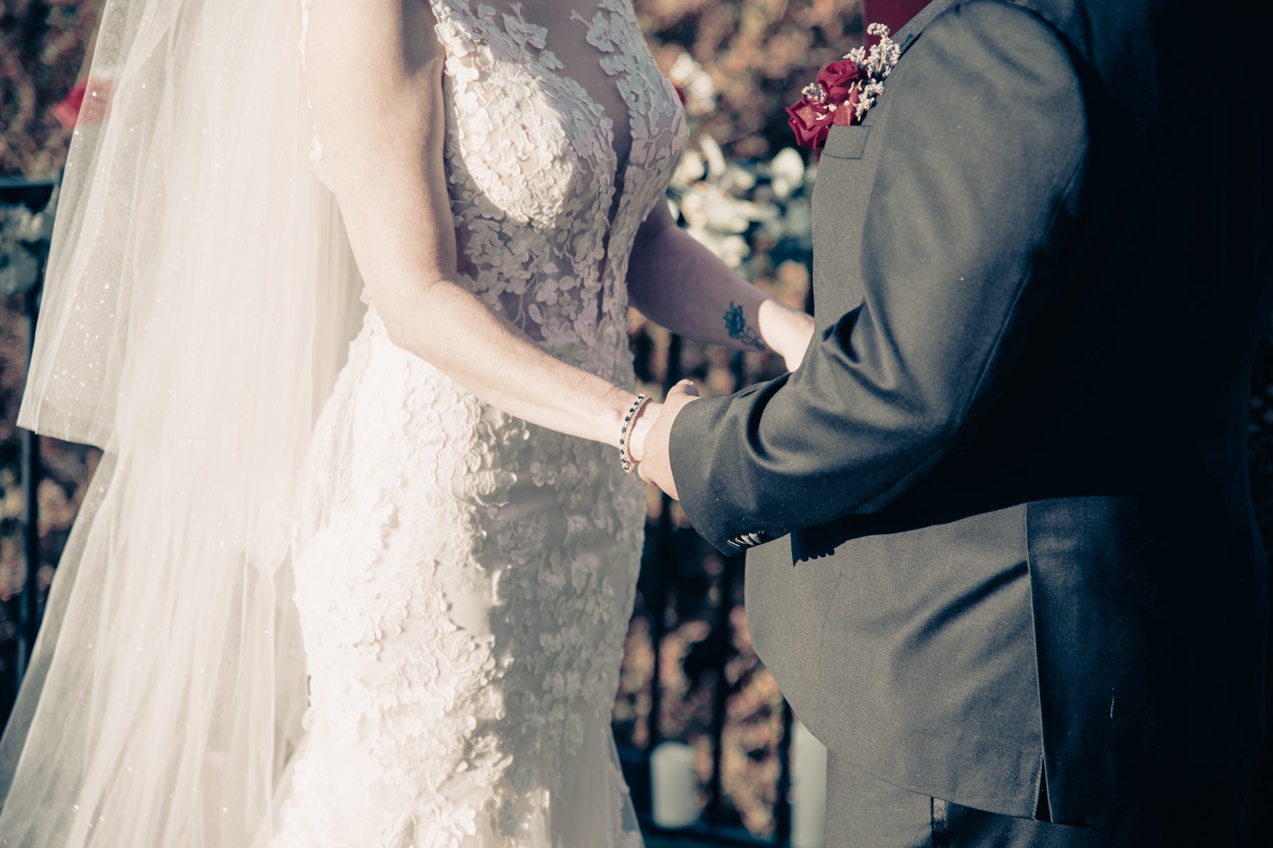 A bride and groom holding hands during their wedding ceremony, with the bride wearing a lace wedding gown and the groom wearing a gray suit with a boutonnière of red and pink flowers.