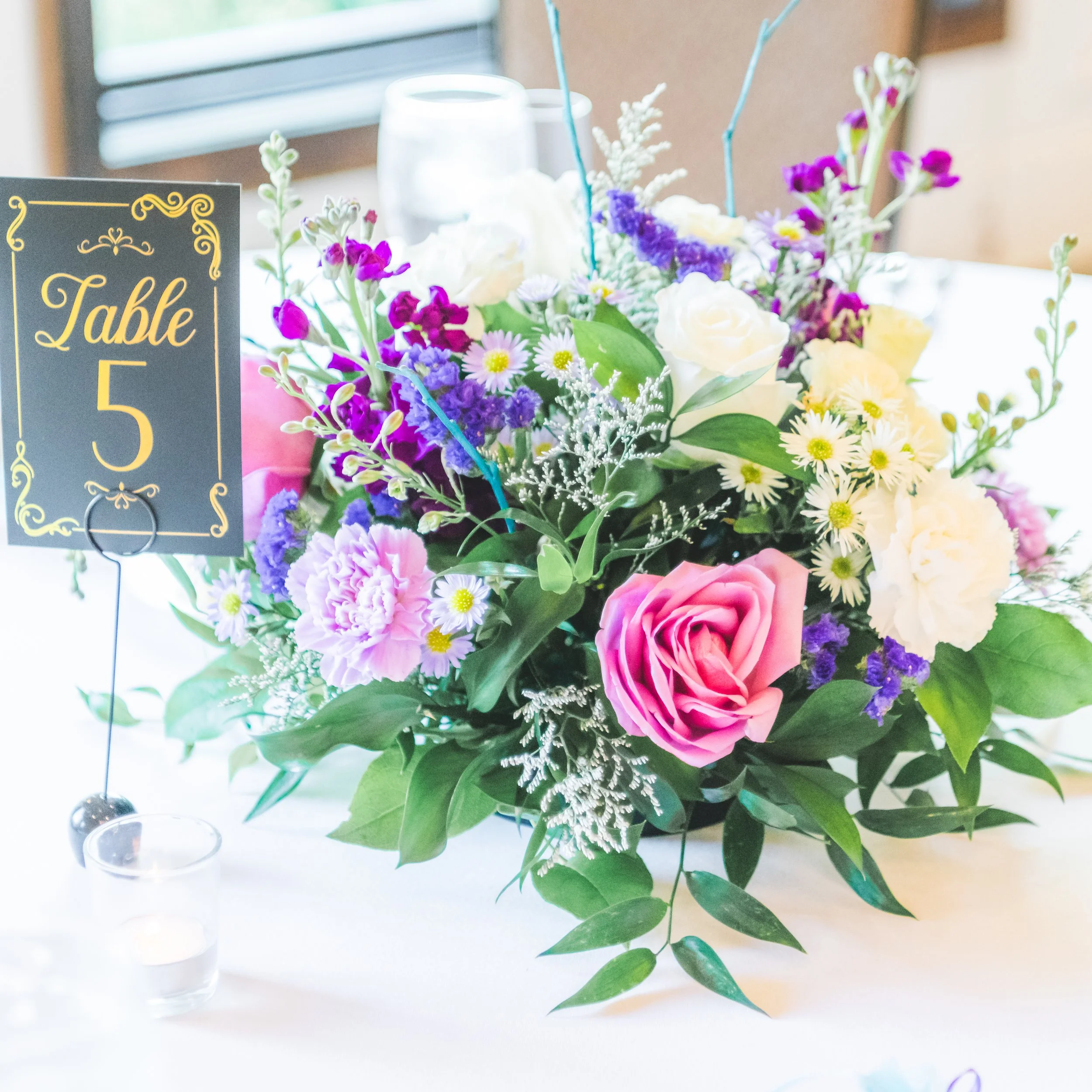 A floral centerpiece with white, purple, pink, and yellow flowers on a table, with a glass of water and a table number card to the left.