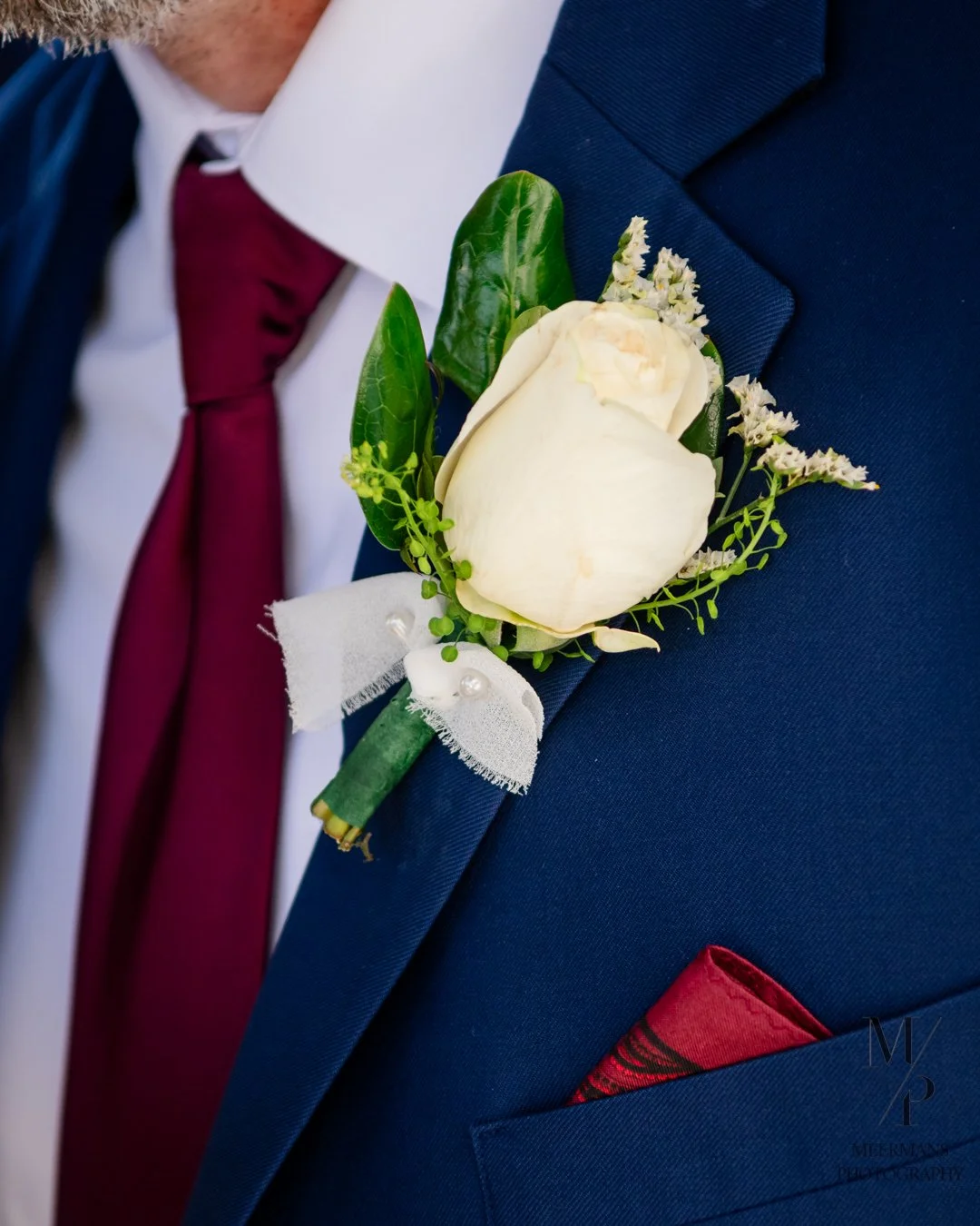 Close-up of a man's suit jacket with a white rose boutonniere, accompanied by green leaves and small white flowers, pinned to the lapel. The man is wearing a dark blue suit, white shirt, and burgundy tie, with a red pocket square visible in the breas