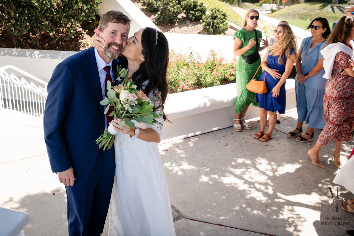 A bride kissing a groom on the cheek at an outdoor wedding, with wedding guests in colorful dresses watching in the background.