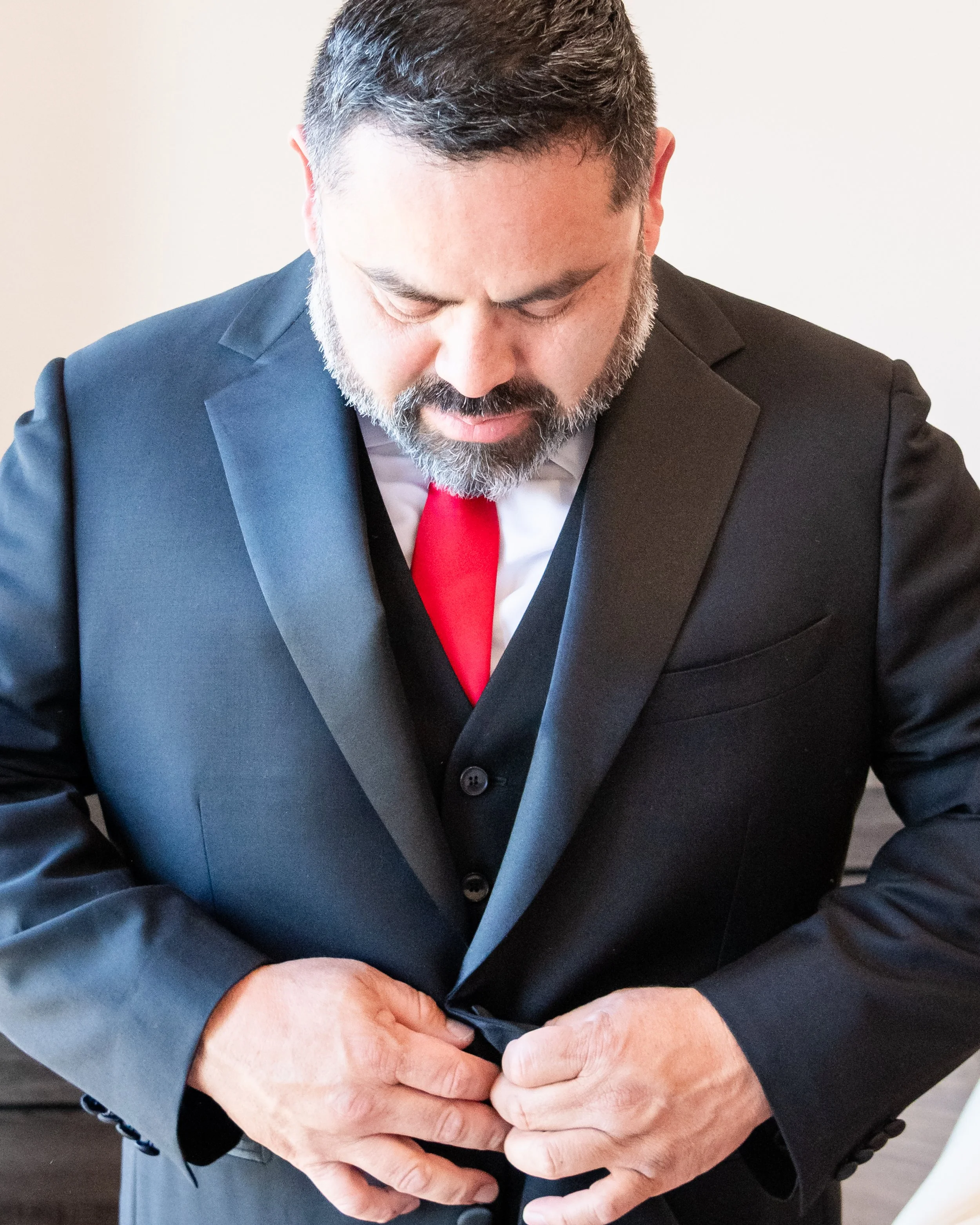 A man in a dark suit and red tie adjusts his suit jacket, standing against a plain light-colored wall.
