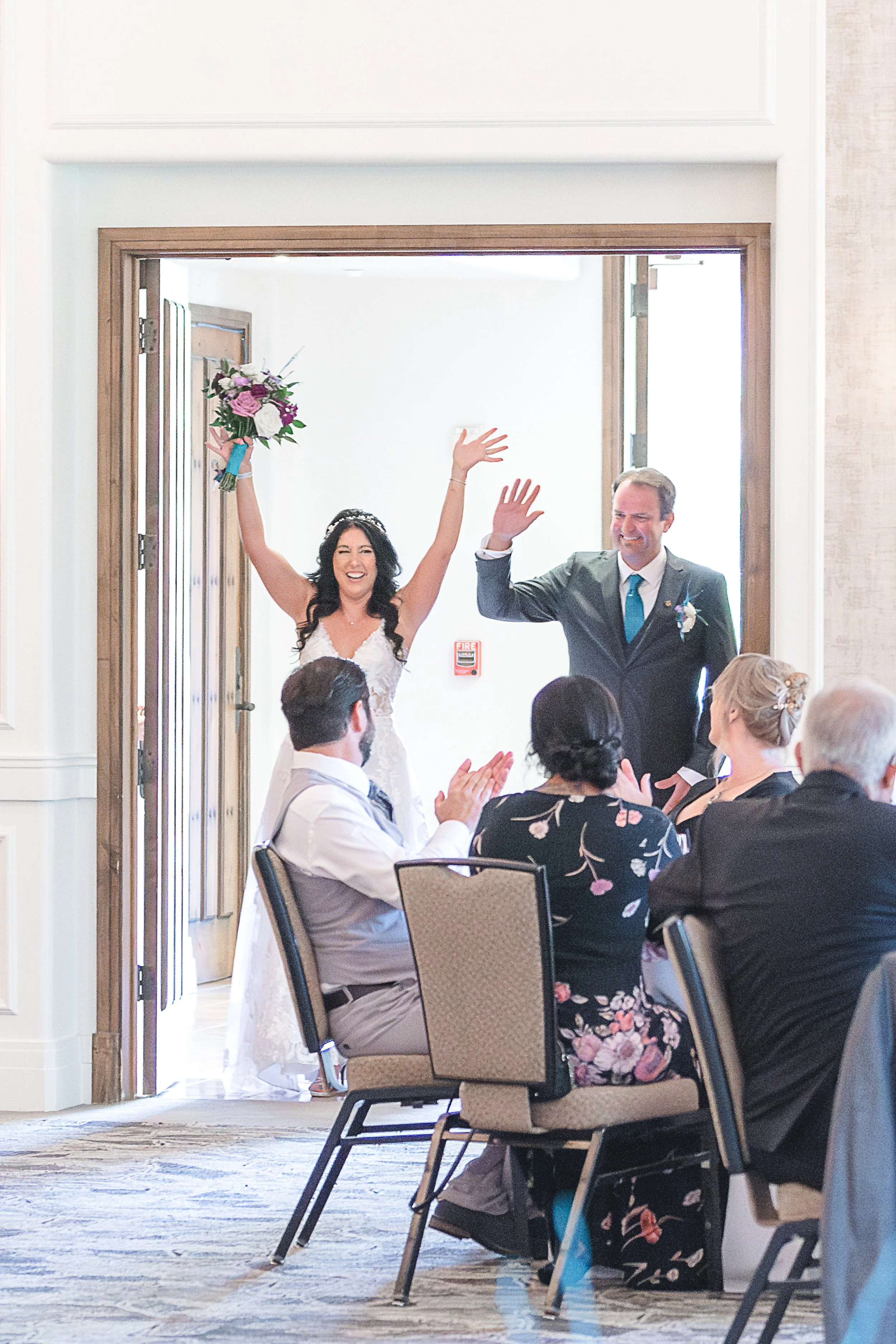 A bride and groom celebrating at their wedding reception, walking into the room with guests clapping, the bride holding a bouquet, and both smiling and waving.