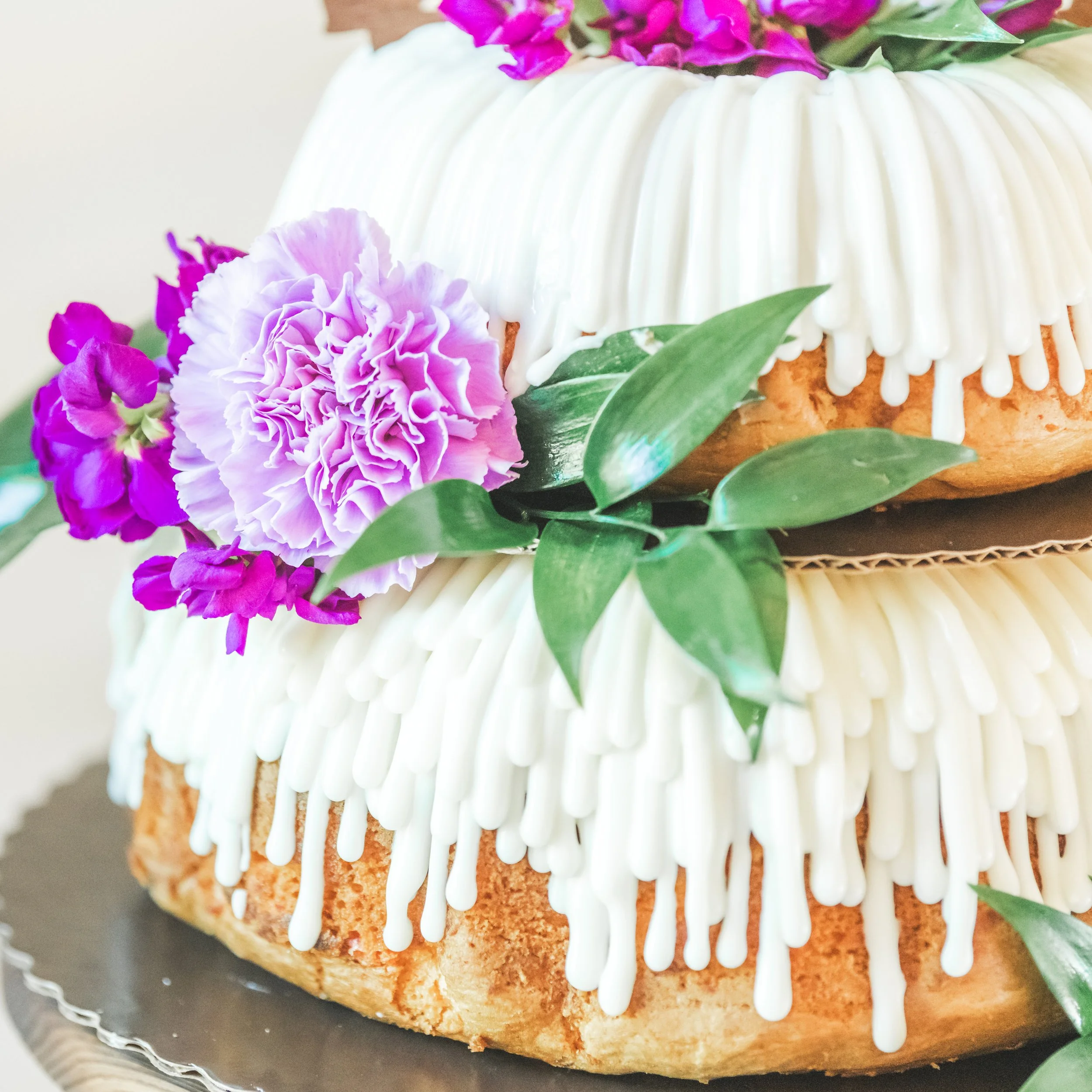 A close-up of a cake with white icing dripping down the sides, decorated with pink and purple flowers and green leaves.