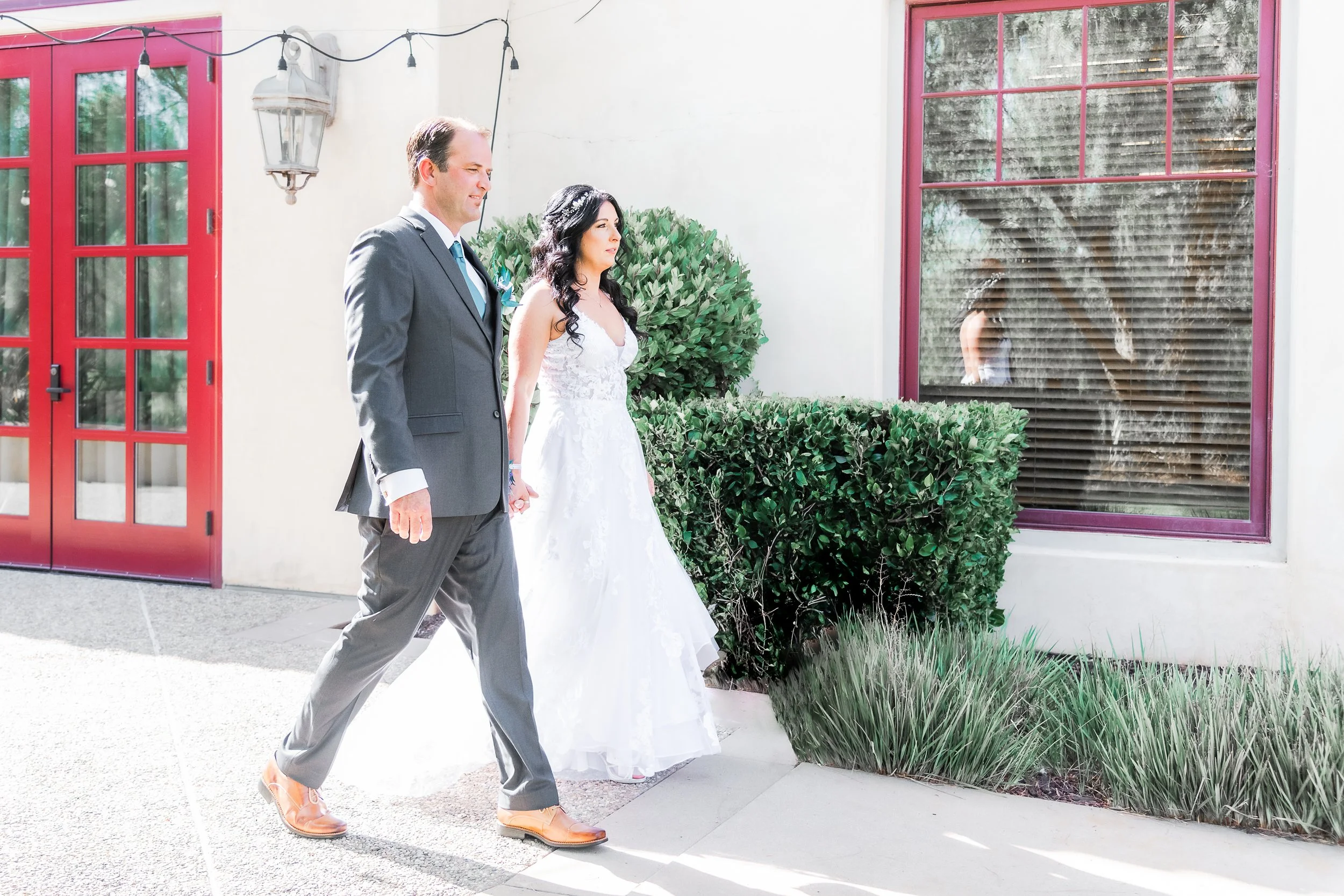 A bride and groom holding hands and walking outside a building with red doors and window frames, surrounded by greenery.