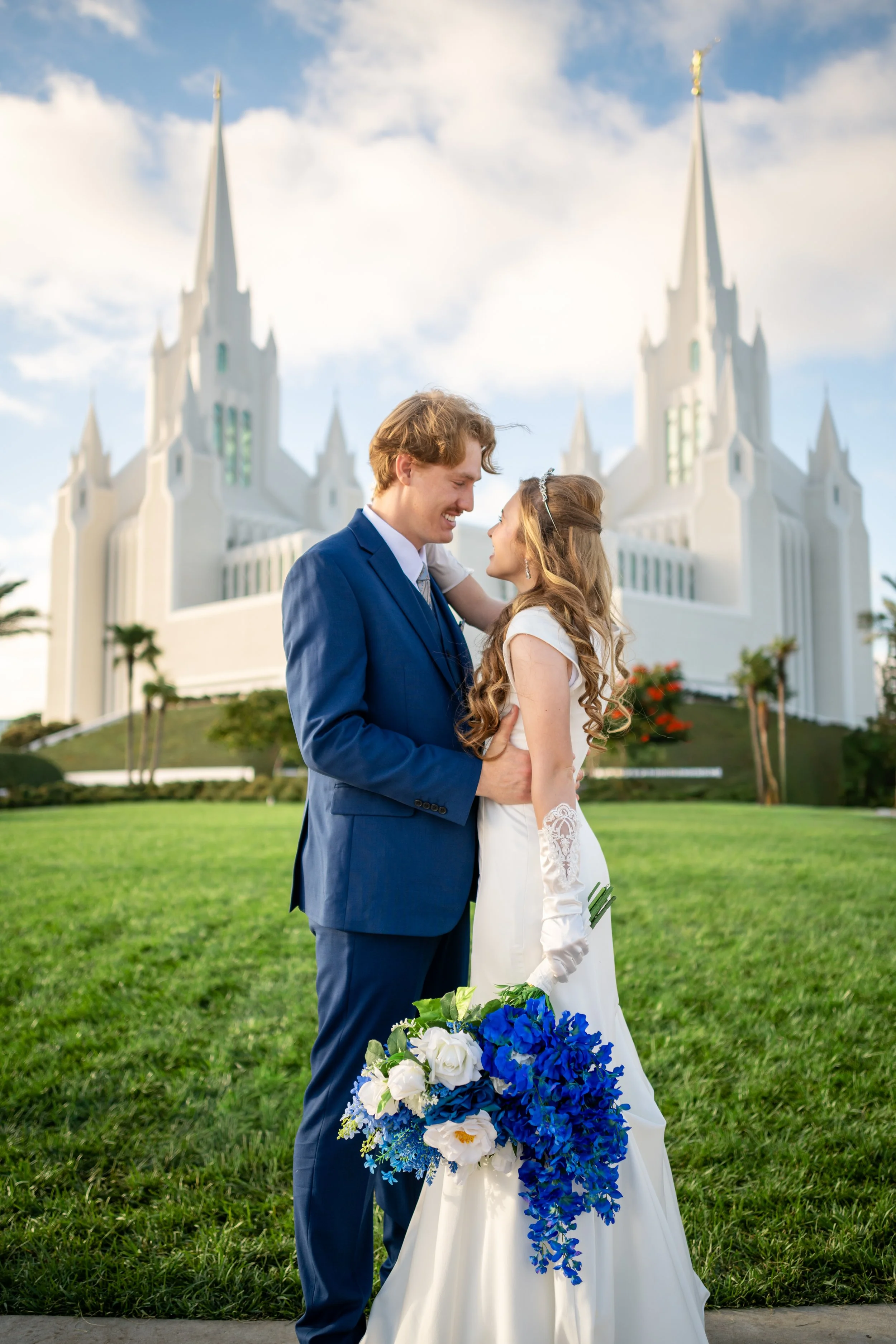 A bride and groom stand close together outdoors in front of a white church with twin spires, smiling at each other, with the bride holding a bouquet of blue and white flowers.