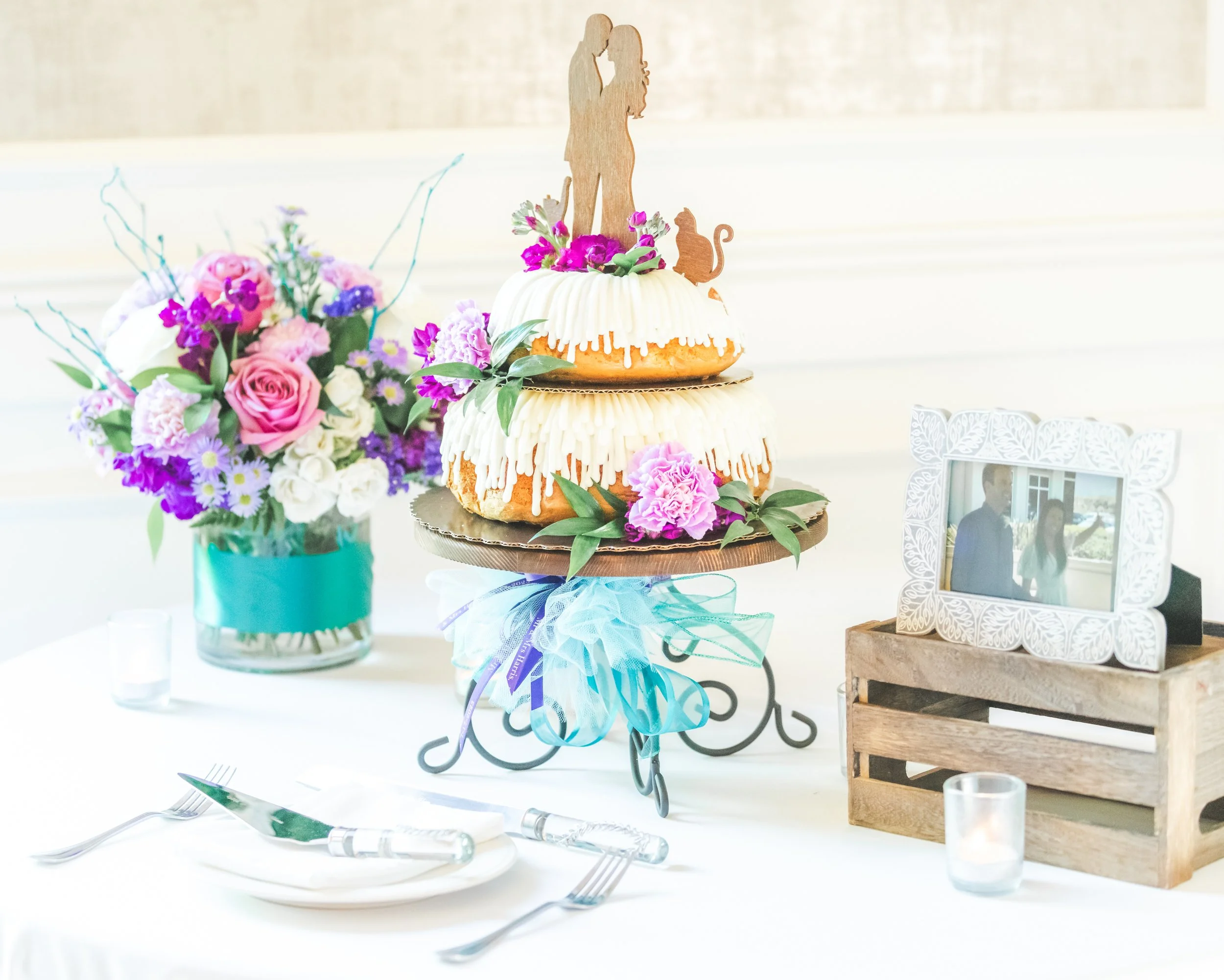 Decorated cake with a wooden topper featuring a couple holding hands, surrounded by pink and purple flowers, placed on a decorative stand on a table with a bouquet of purple, pink, and white flowers nearby.