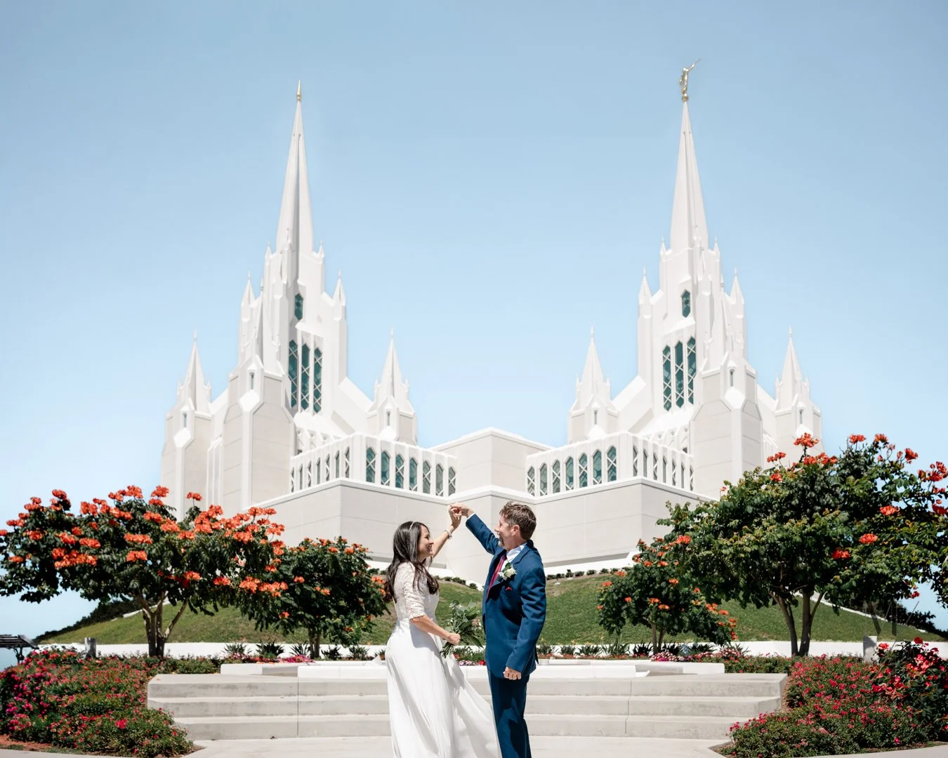 A newlywed couple in wedding attire dancing outside the Salt Lake Temple with colorful flowers and trees in the foreground.