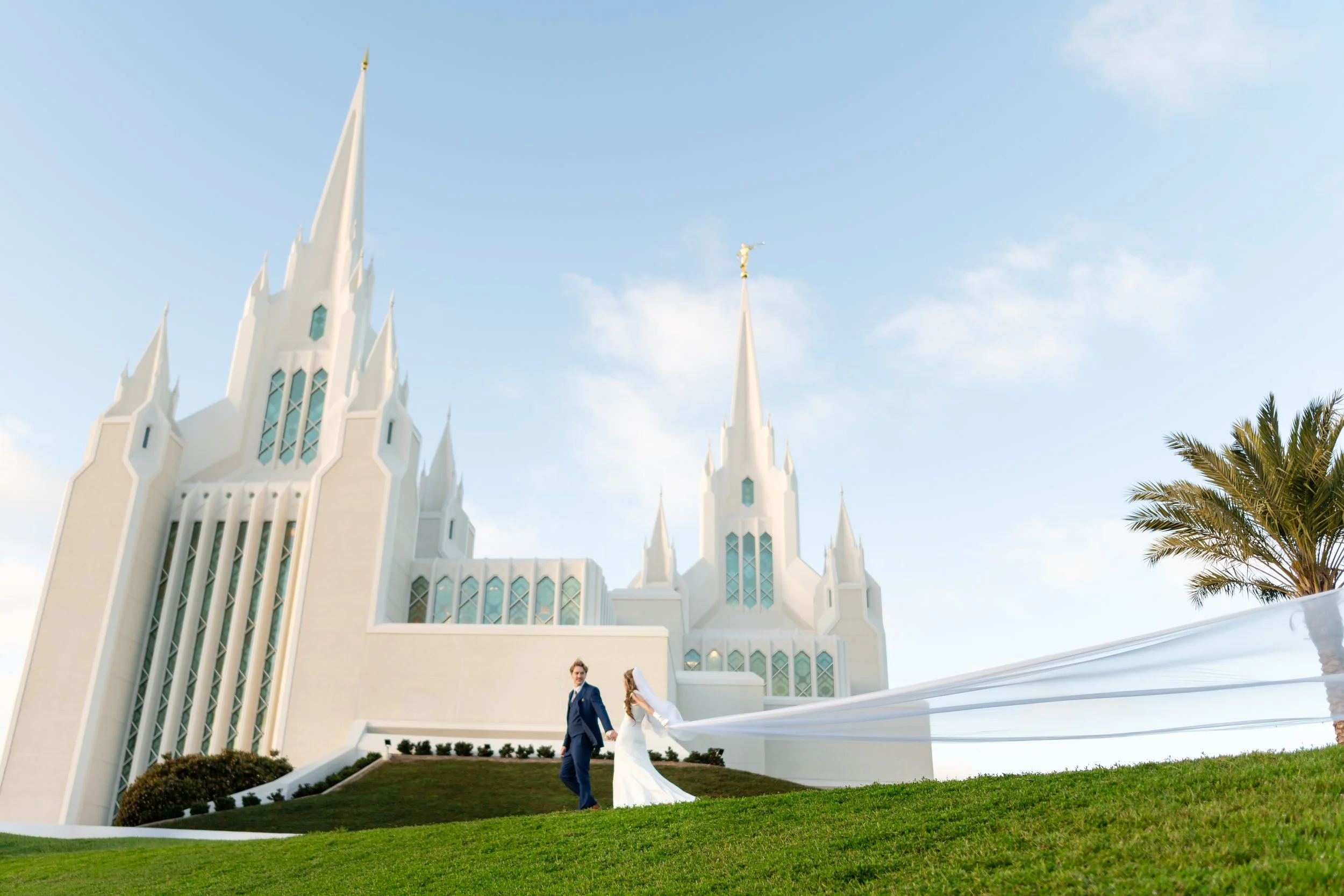 A bride and groom holding hands and walking outside a white church with tall spires and stained glass windows, with a blue sky and palm trees in the background.