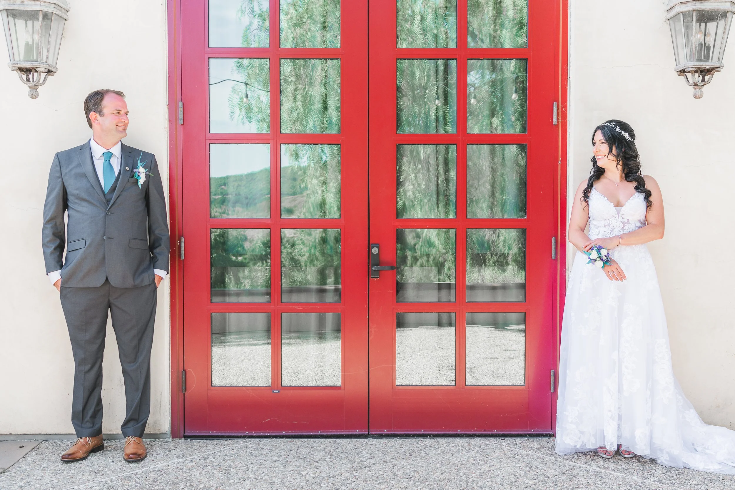 A bride and groom stand apart on either side of a red door, smiling at each other. The groom is wearing a gray suit with a blue tie and boutonniere, while the bride is wearing a white wedding gown and holding a bouquet, with a floral headpiece.