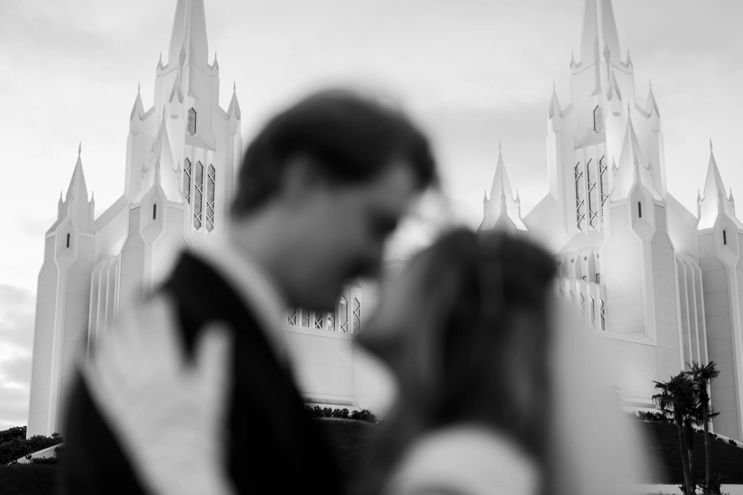 Blurry silhouette of two people kissing in front of a white church with spires surrounded by palm trees.