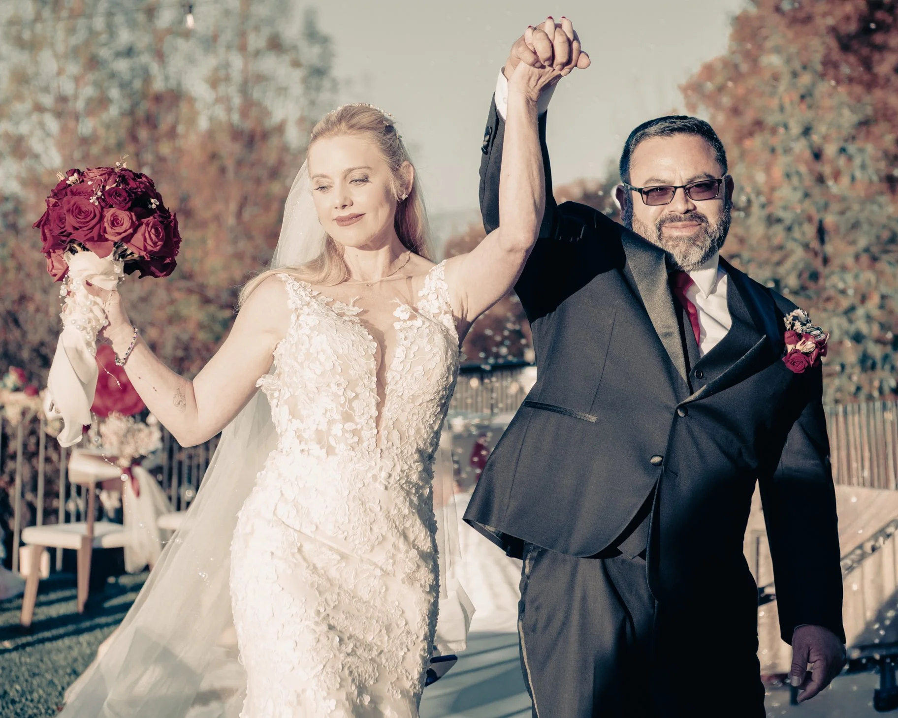 A bride and groom holding hands high after their wedding, outdoors with trees and a railing in the background.