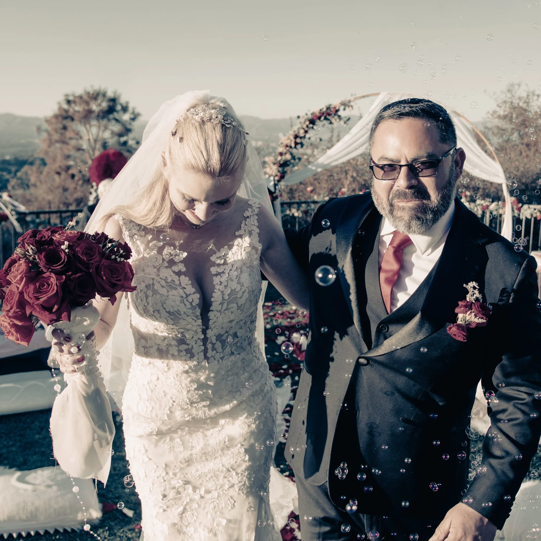 A bride and groom, dressed in wedding attire, celebrating at their outdoor wedding ceremony with bubbles in the air and scenic landscape in the background.
