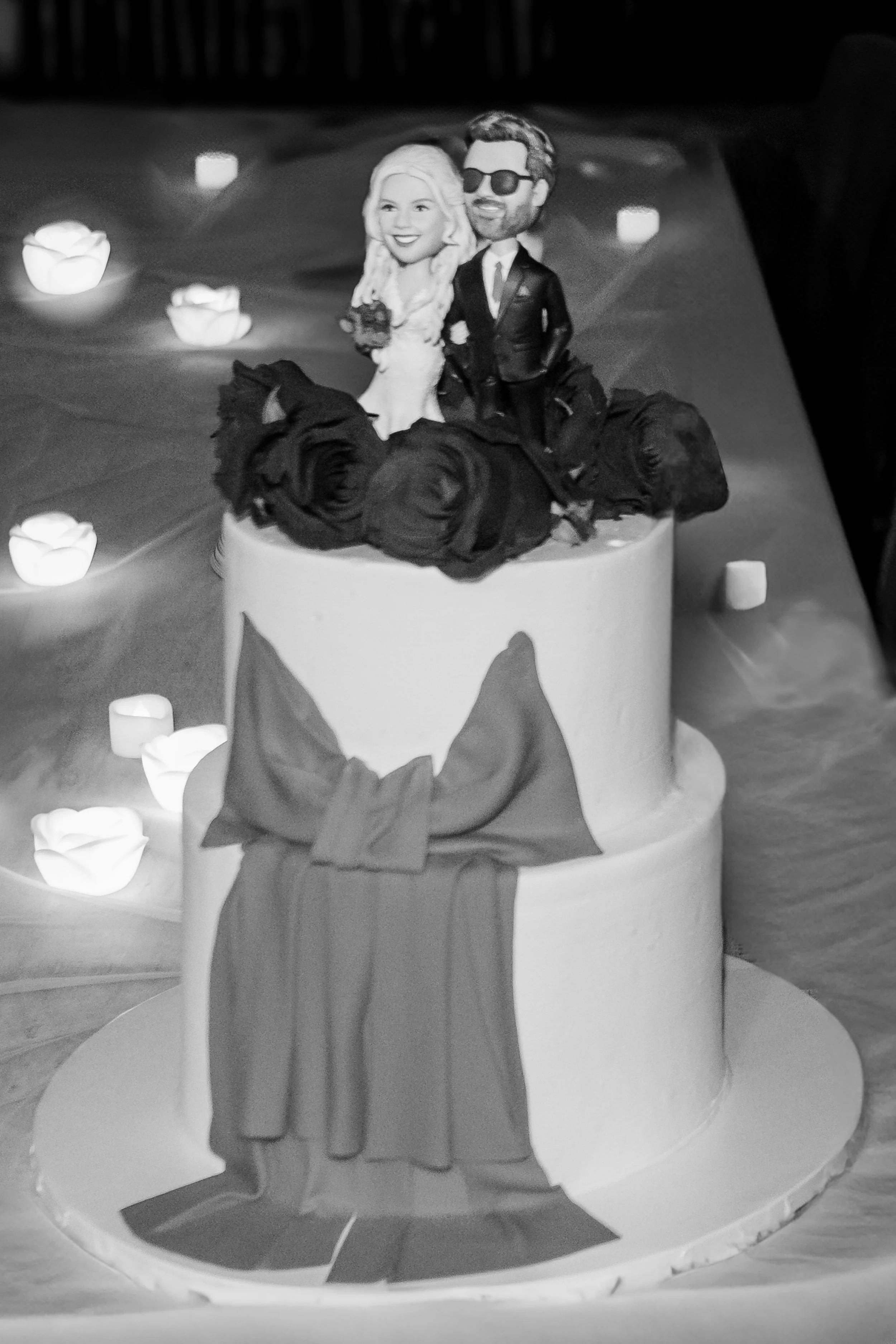 Cake with cartoon topper of a smiling woman in white dress and a man in sunglasses and a suit, surrounded by decorative roses and candles.