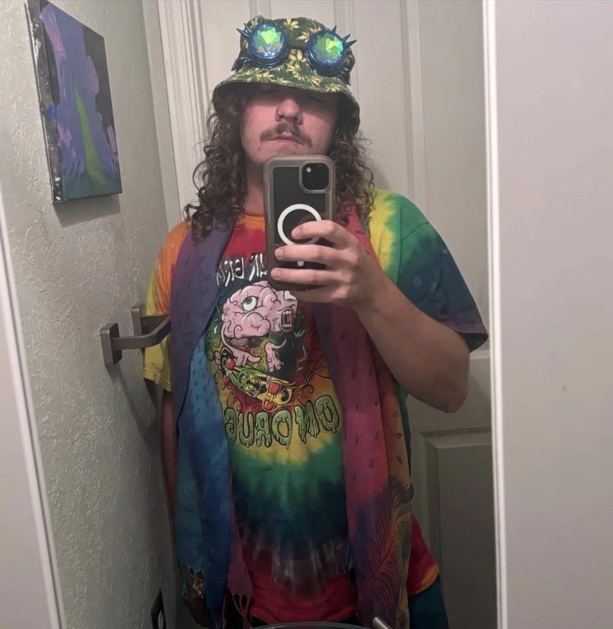 Person taking a mirror selfie wearing a tie-dye shirt with a cartoon brain and the words 'DUMB DUMB', colorful rainbow-colored scarf, a bucket hat with spiked goggles, and curly hair.