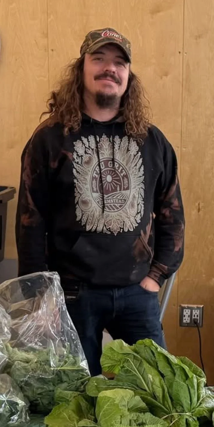 A man with long curly hair, a beard, wearing a camouflage cap and a black hoodie with a graphic design, stands in front of a wooden wall. In front of him are various leafy green vegetables on a table, some in clear plastic bags.