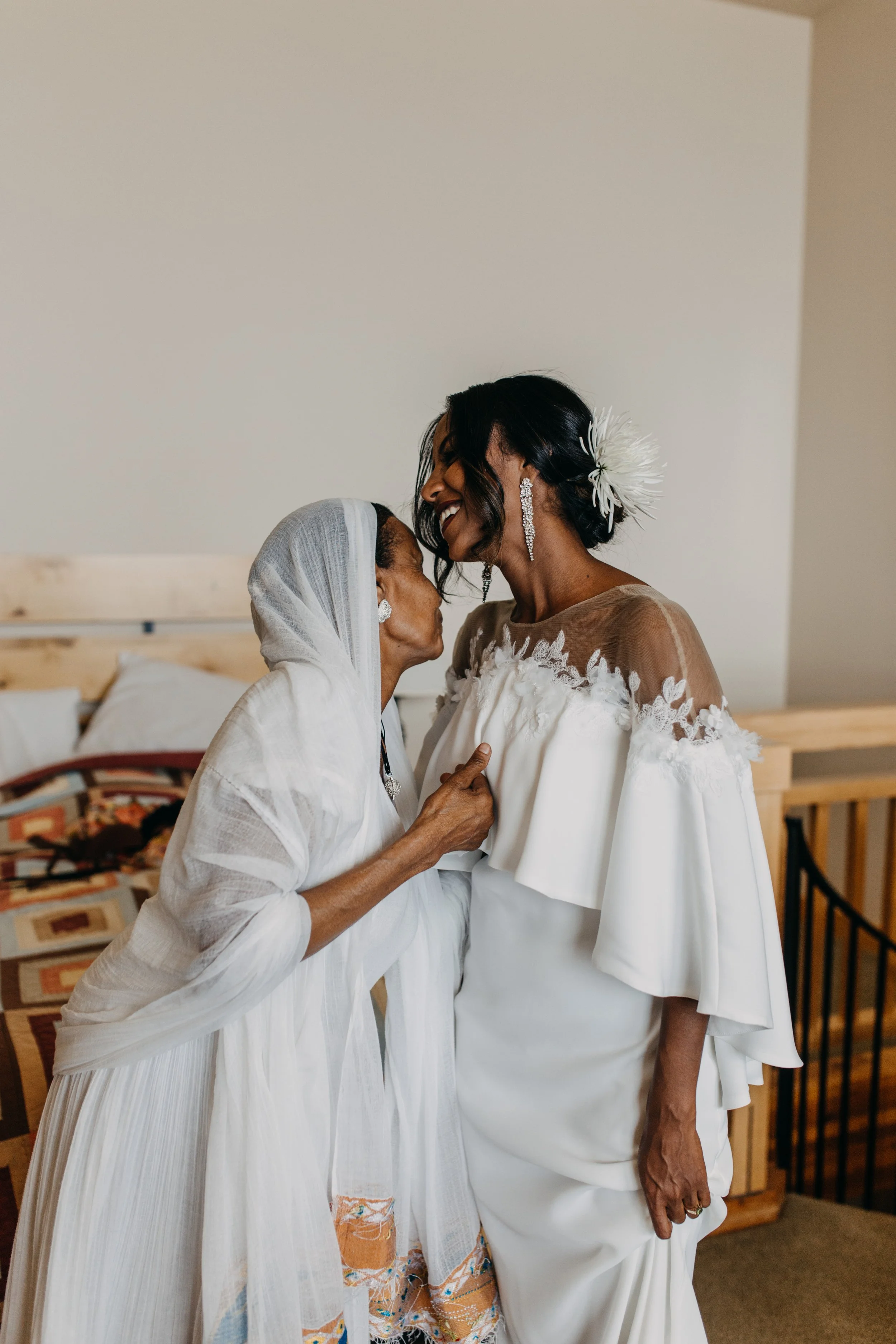 An elderly woman in traditional Ethiopian attire sharing a joyful moment with a bride in a white dress, both smiling and embracing each other indoors.