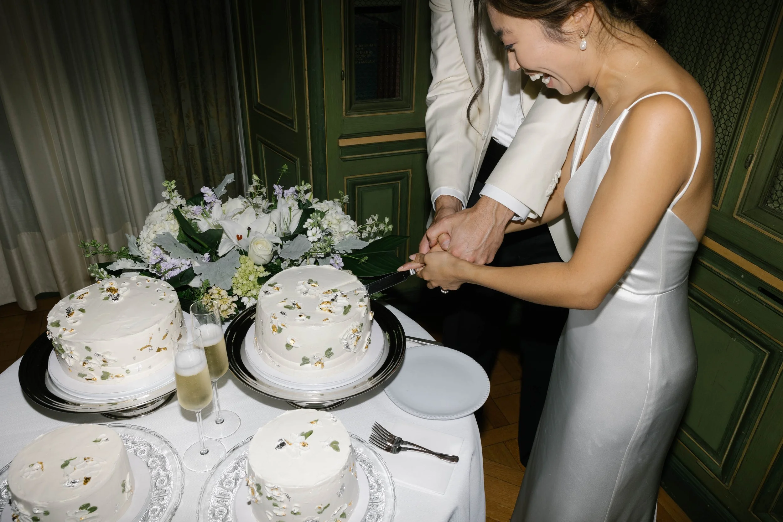 A couple cutting a wedding cake together, dressed in formal attire, surrounded by flowers and champagne glasses on a table.