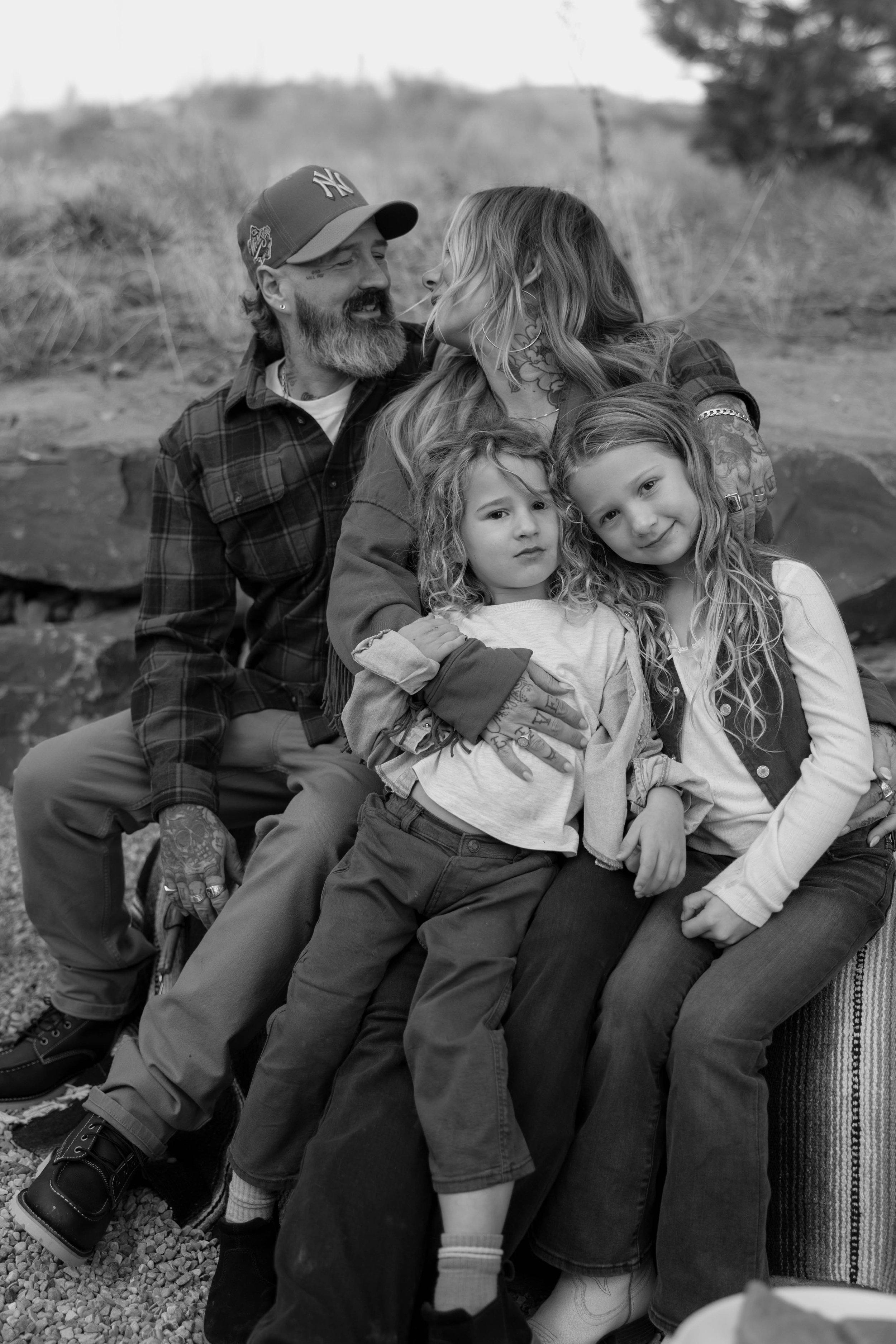 A black and white photo of a family sitting outdoors. A bearded man wearing a baseball cap sits next to a woman with long hair. Two young children are sitting on their laps. The background is blurred nature.